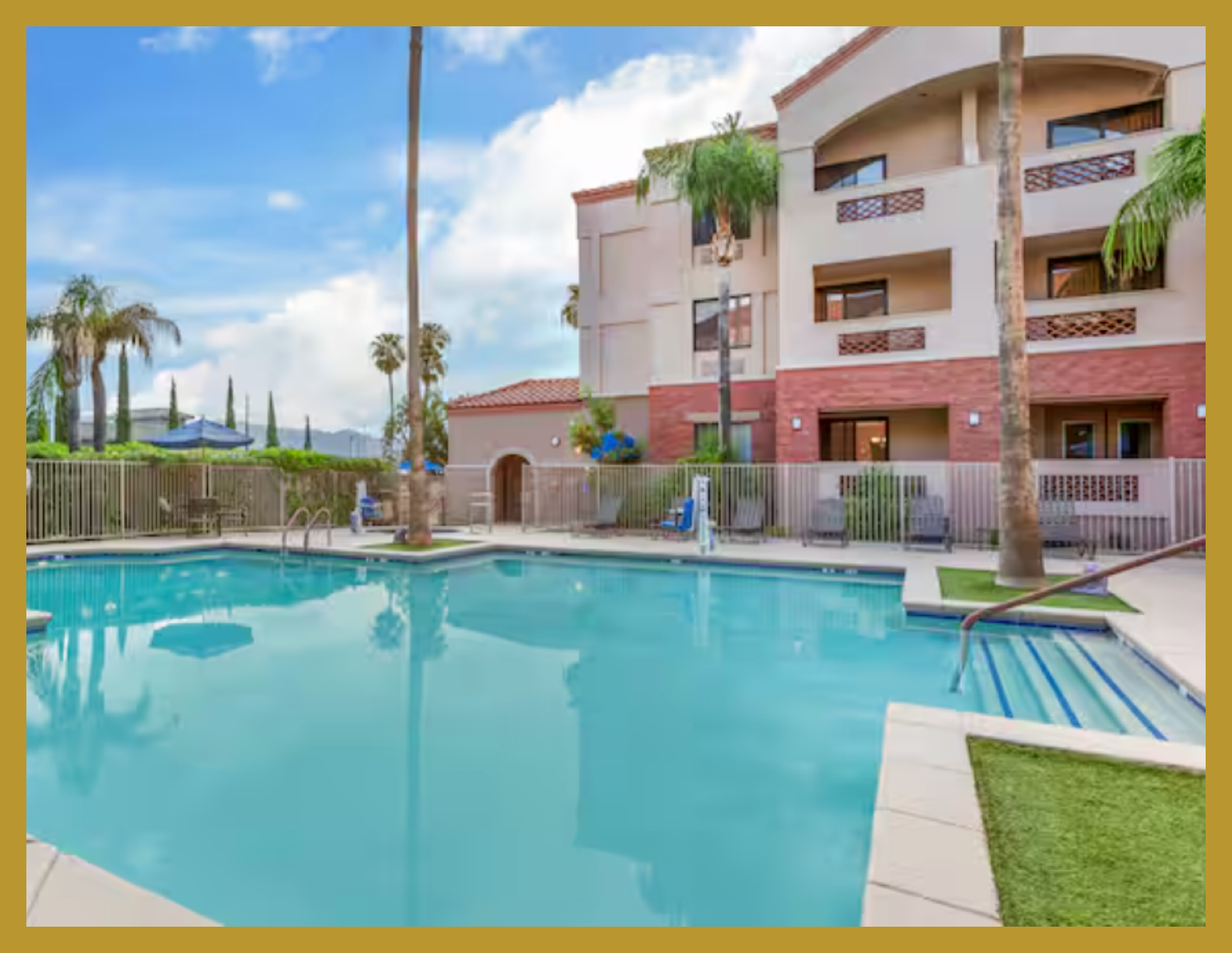 Outdoor swimming pool with ladder and surrounding lounge chairs, palm trees, apartment building with balconies, fence, and cloudy sky in the background.