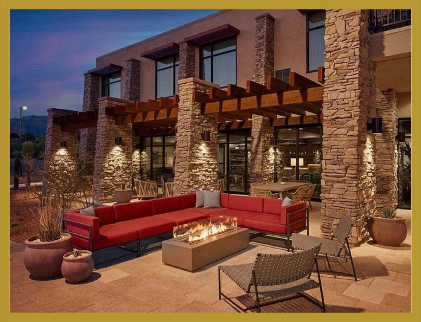 Outdoor patio area with red sectional sofa, fire pit, and patio chairs in front of a building with stone pillars and large windows, during evening.