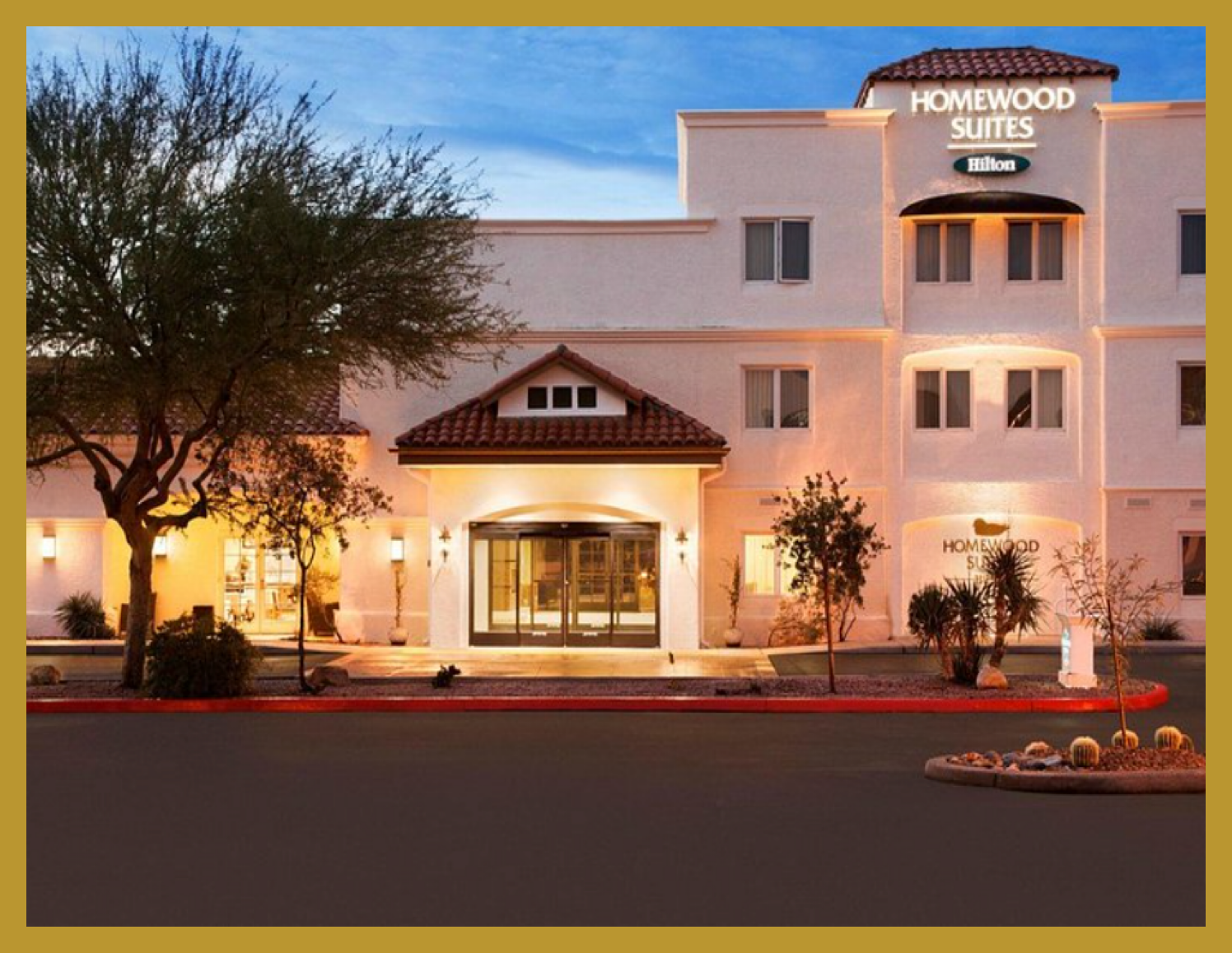 Exterior of a Homewood Suites hotel at dusk with warm lighting, white stucco walls, and a red-tiled roof, trees, and desert landscaping in front.