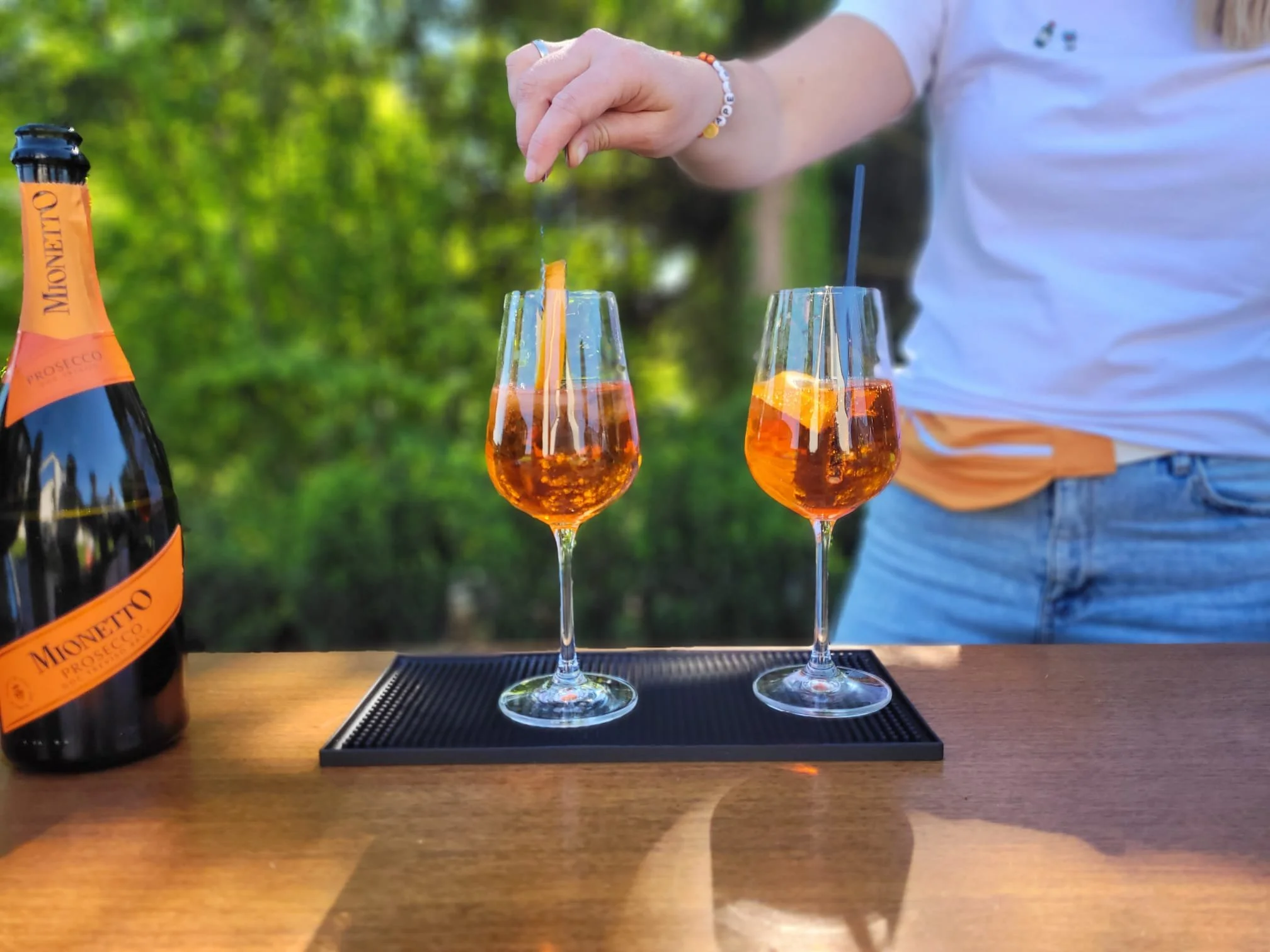 Zwei Gläser mit orangefarbenem Getränk, ein Sekt oder Wein, auf einem schwarzen Teller, mit Flasche Mionetto Prosecco links im Bild, draußen mit grünen Bäumen im Hintergrund.