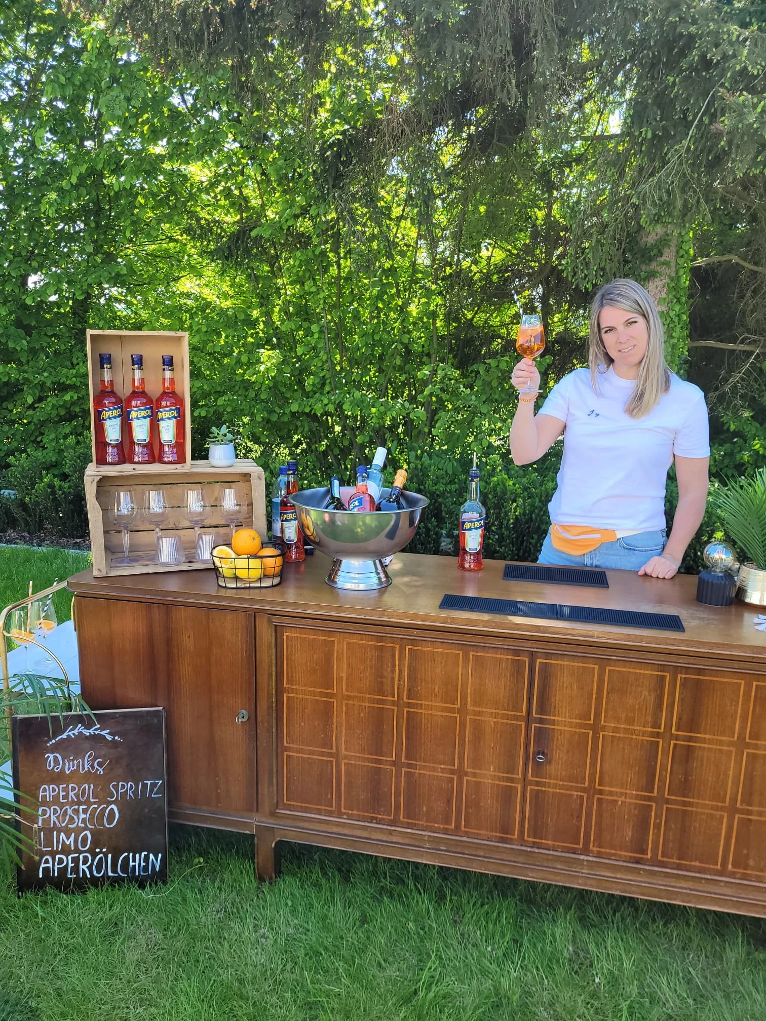Eine Frau steht hinter einem Barwagen im Garten, hält ein rosafarbenes Getränk in der Hand und lächelt. Der Barwagen ist mit Aperol-Getränken, Gläsern, Zitrusfrüchten und einer kleinen Tafel mit Getränkeliste dekoriert. Im Hintergrund sind Bäume und Büsche.