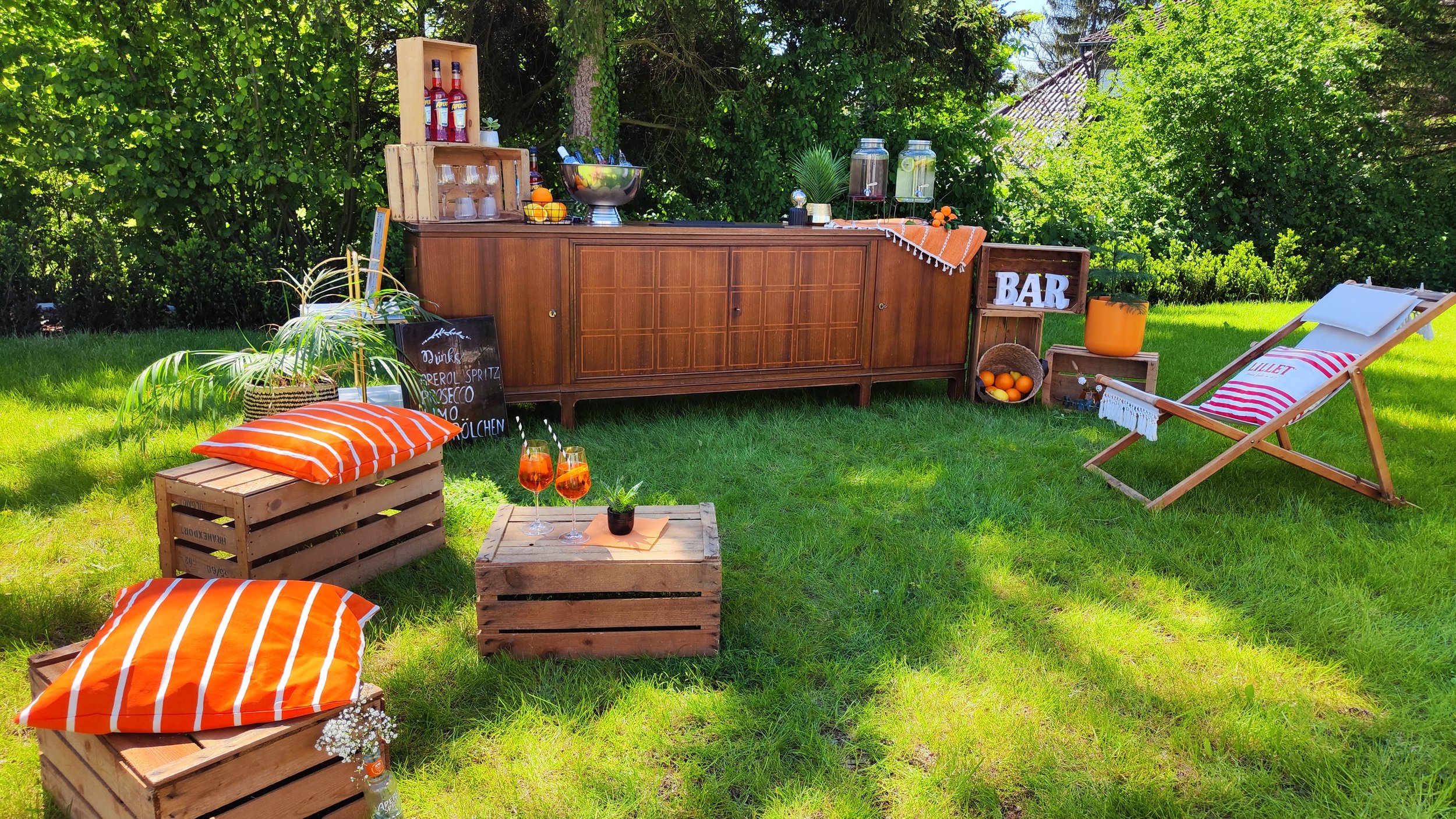Ein Gartentisch mit Getränken, eine Bar im Freien, Holzstühle mit Kissen, Pflanzen und Sonnenschirme, sonniges grünes Gartenumfeld.