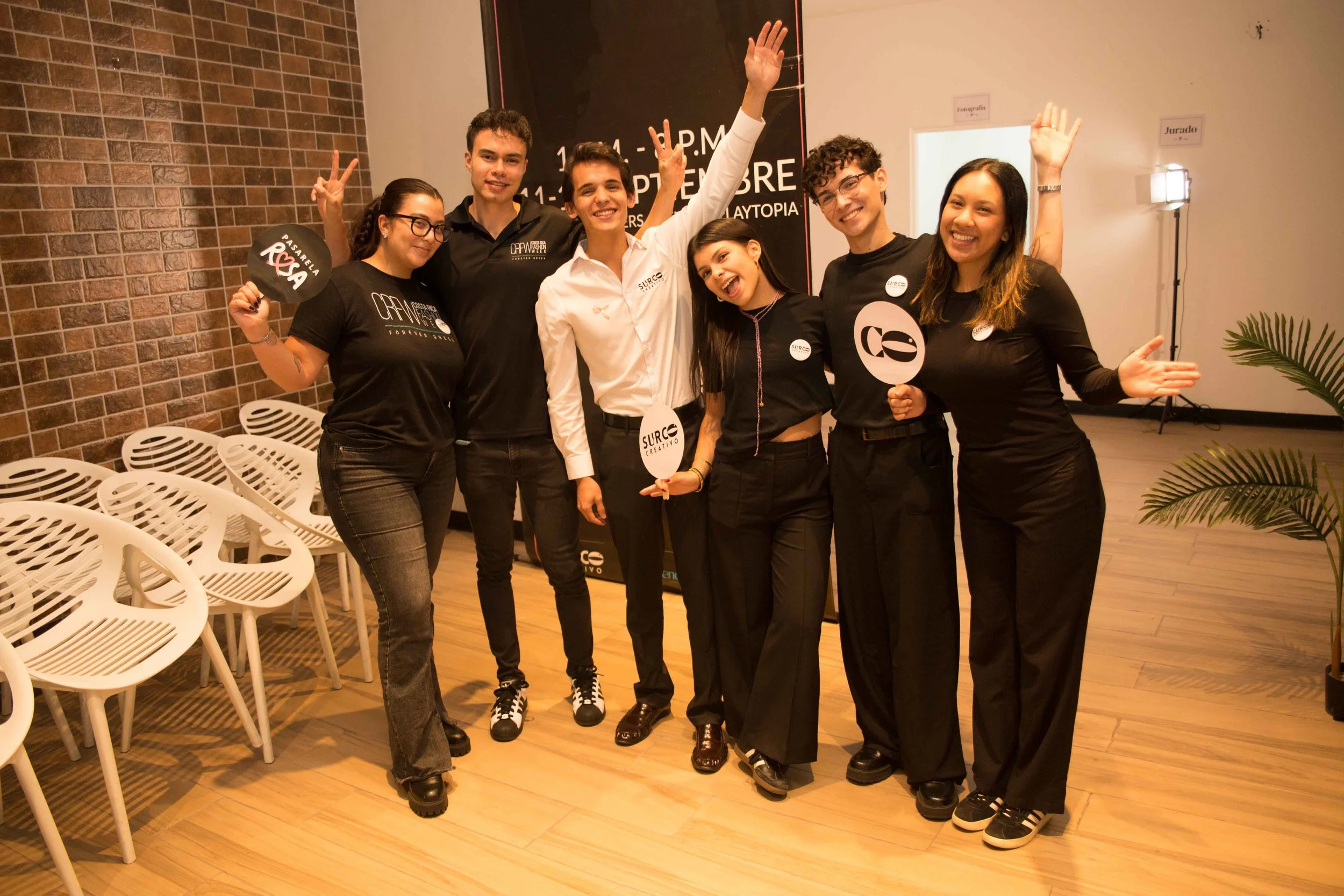 Grupo de siete personas posando y sonriendo en un evento, algunos sosteniendo carteles con nombres o logotipos, fondo con cartel y sillas blancas apiladas.