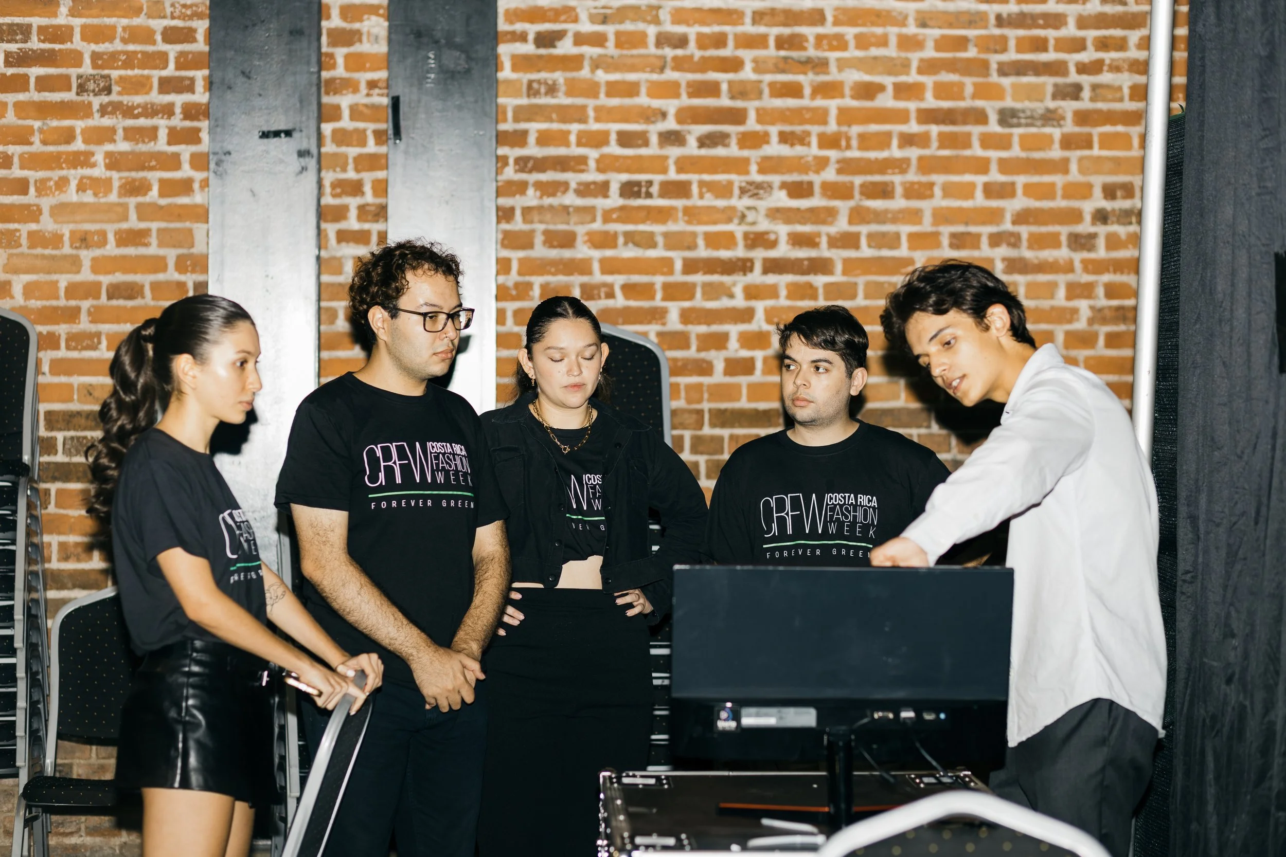 Un grupo de cinco personas, cuatro usando camisetas negras con el logo de CREW Costa Rica Fashion Week, están en una sala con paredes de ladrillo y metal. La persona de la derecha, que lleva una camisa blanca, parece estar mostrando algo a los demás.