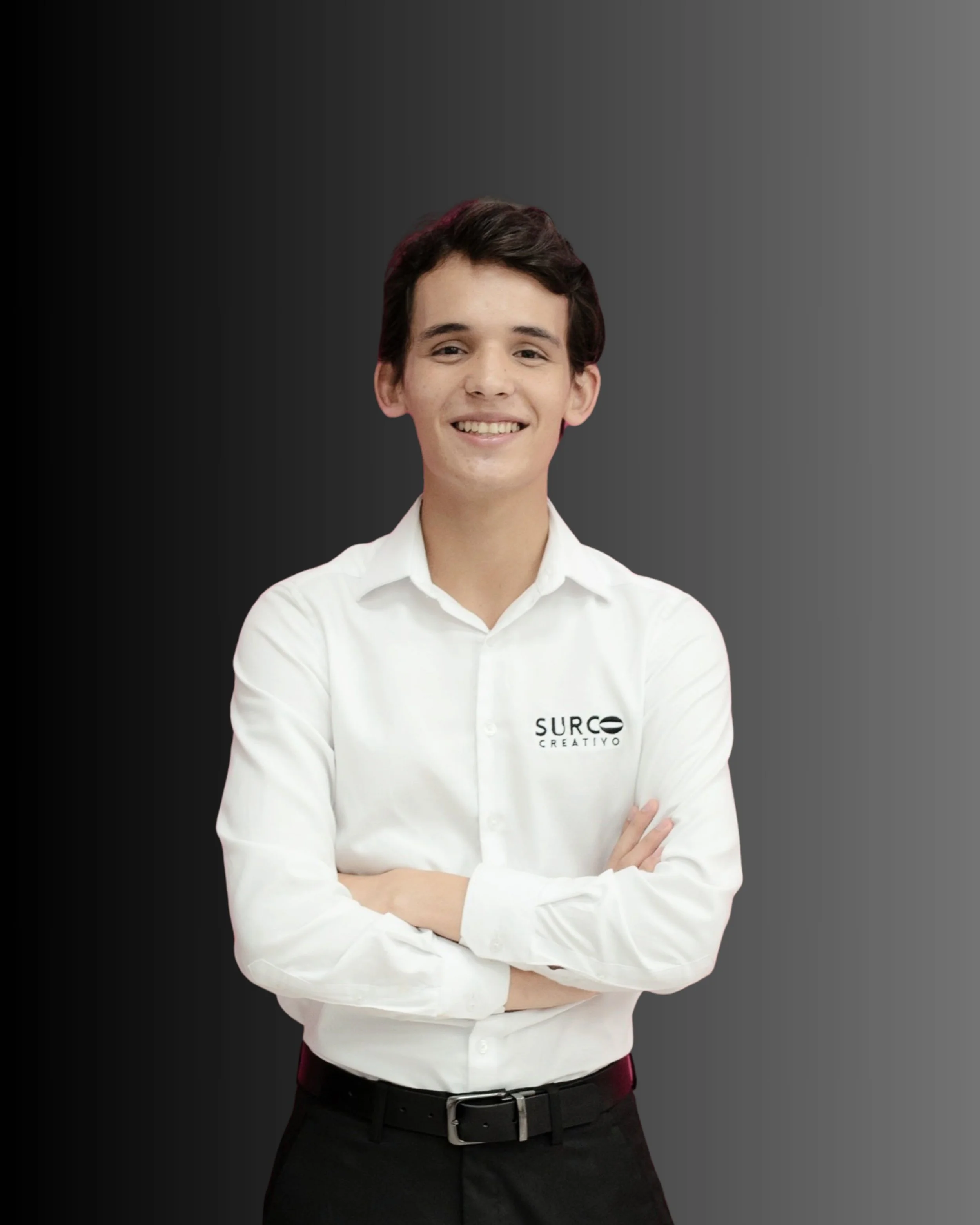 Joven sonriendo con brazos cruzados, lleva camisa blanca con logo de 'Surco Creativo' y fondo gris oscuro.