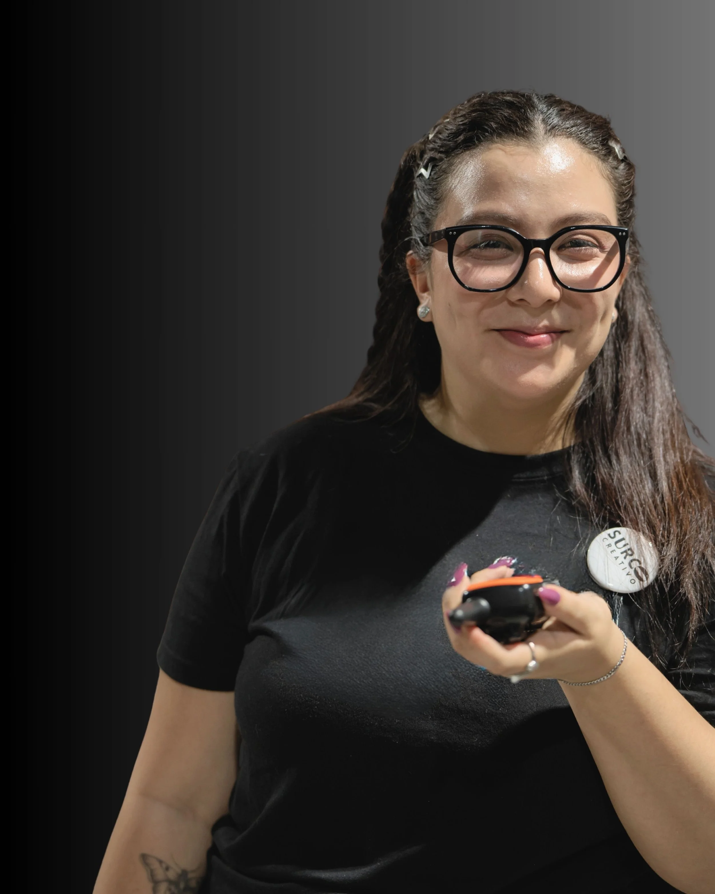 Mujer sonriendo con gafas negras, usando una camiseta negra y sosteniendo un teléfono móvil, con un botón de campaña en su ropa que dice 'Sucre Creativo'.