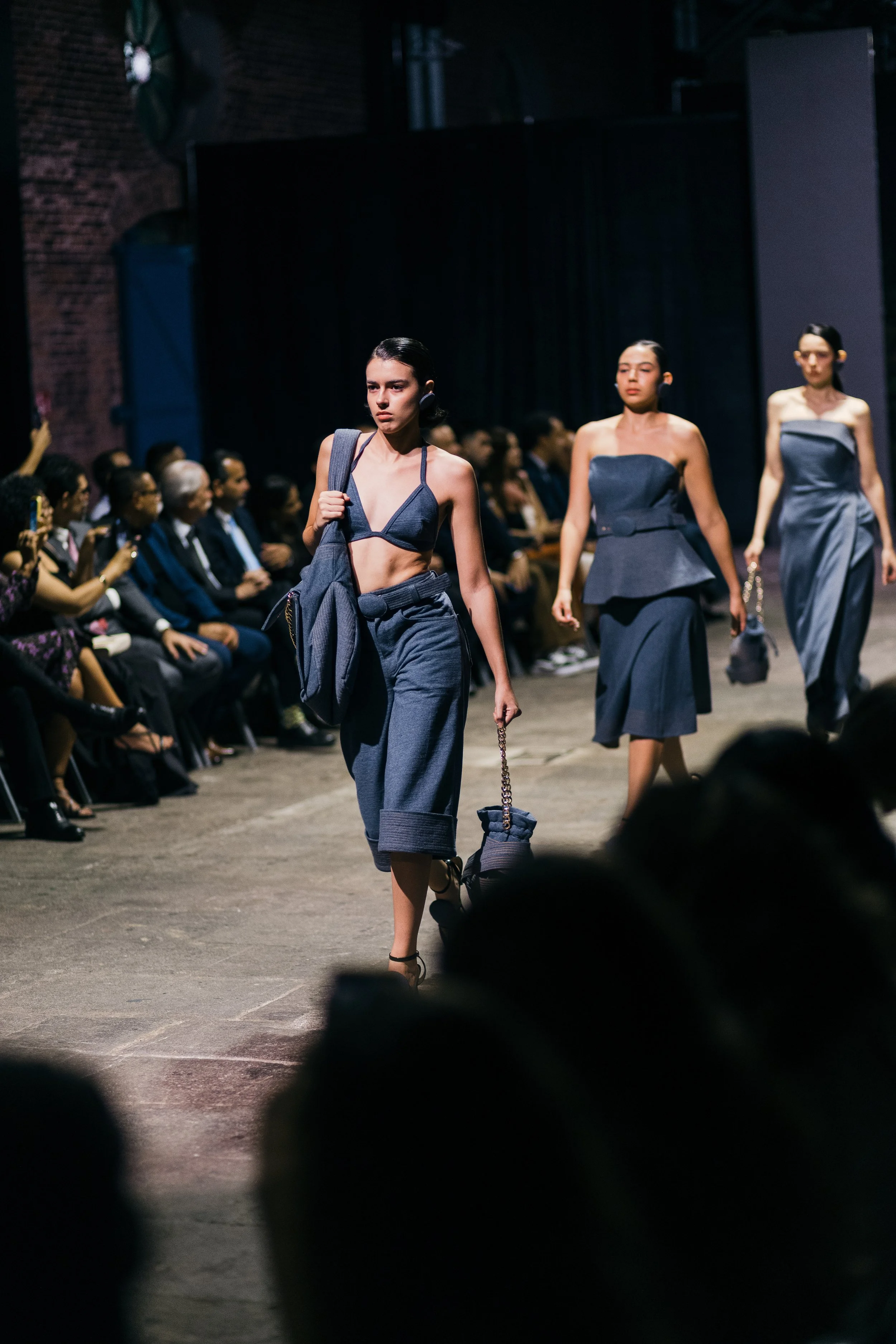 Modelos desfilando en una pasarela con ropa de color azul oscuro, mientras la audiencia observa.