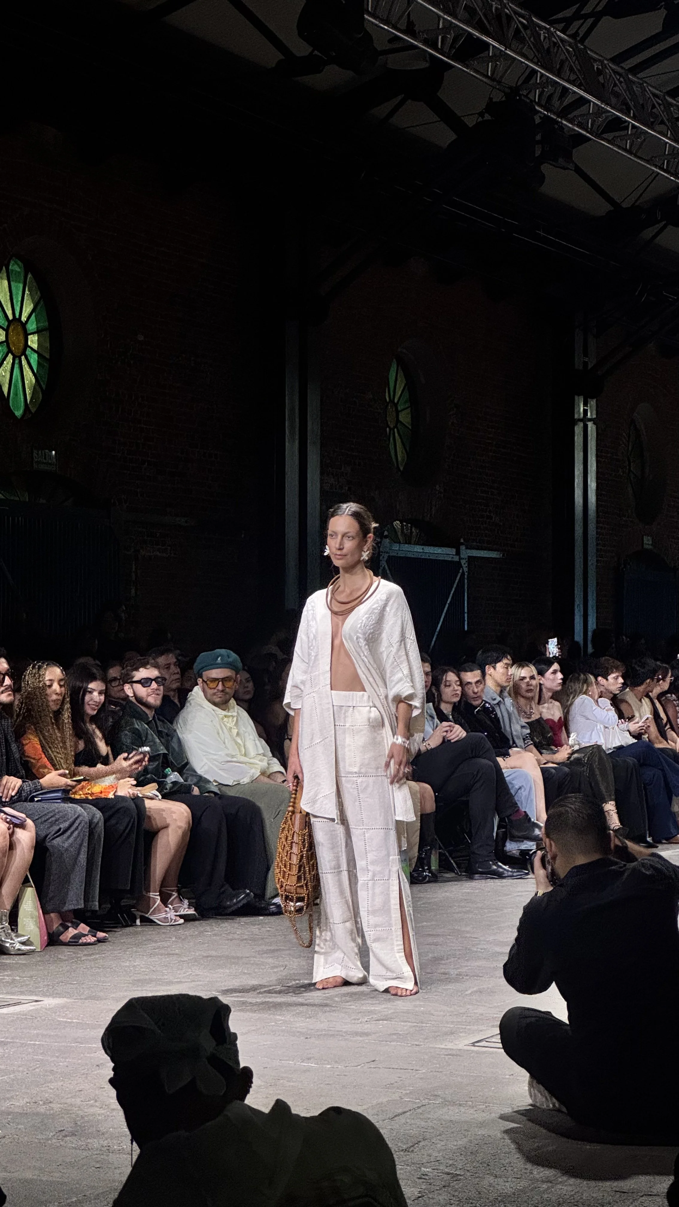 Modelo de moda en un desfile, usando ropa blanca, en un escenario con público sentado a los lados y ventanales de estilo antiguo en la pared de fondo.