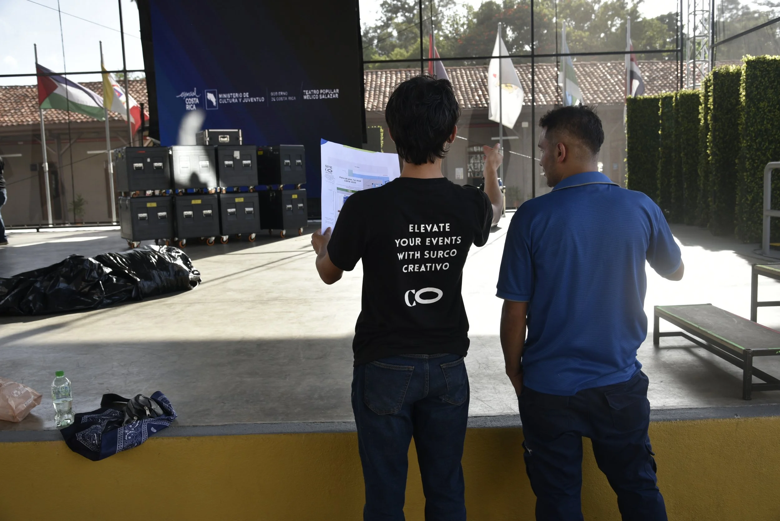 Dos personas viendo un escenario preparado, con banderas en el fondo, en un espacio cubierto con ventanas grandes y una pared decorada con plantas.
