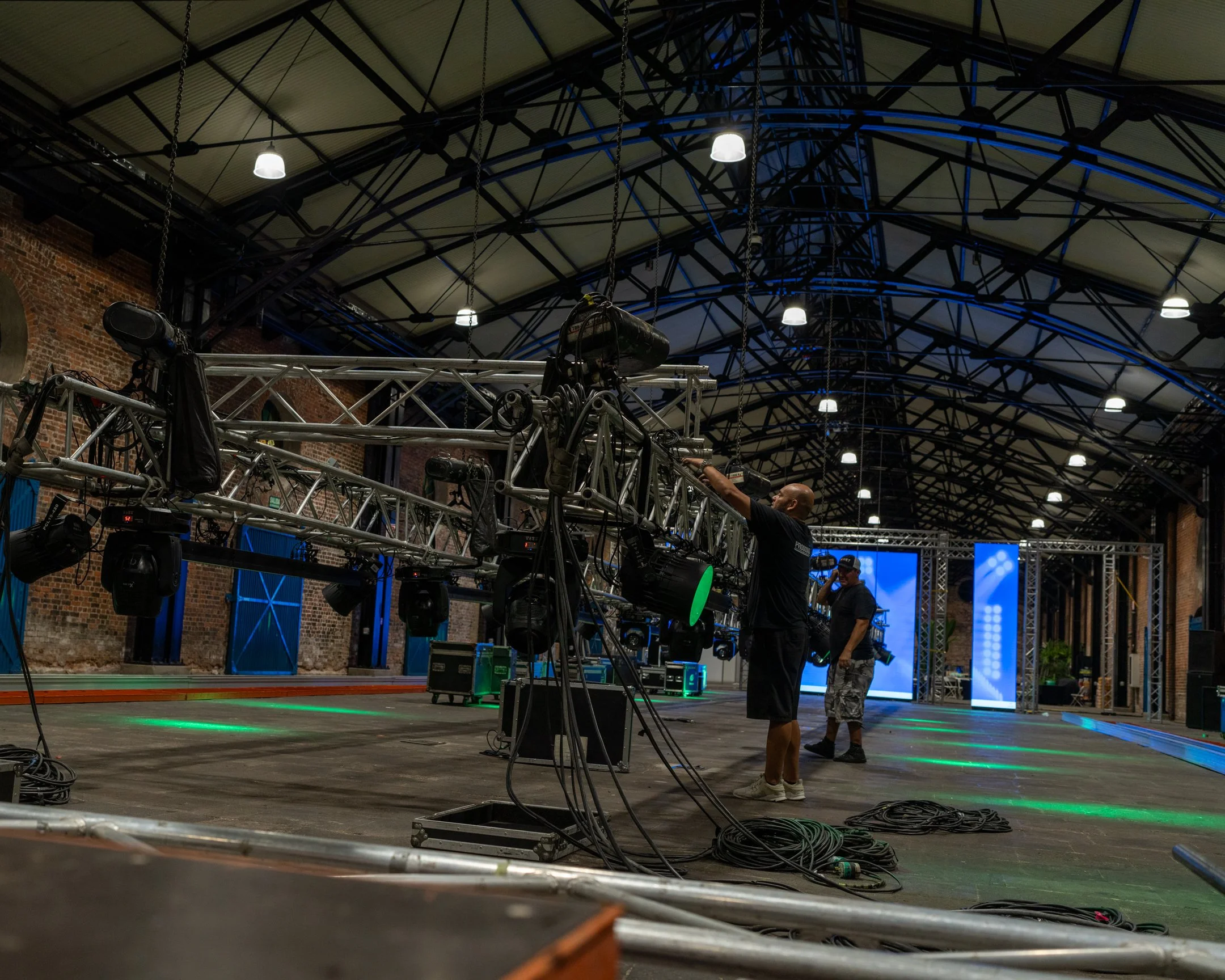 Personal técnico ajustando equipo de iluminación y sonido en un escenario techado, con paredes de ladrillo y estructura metálica visible.