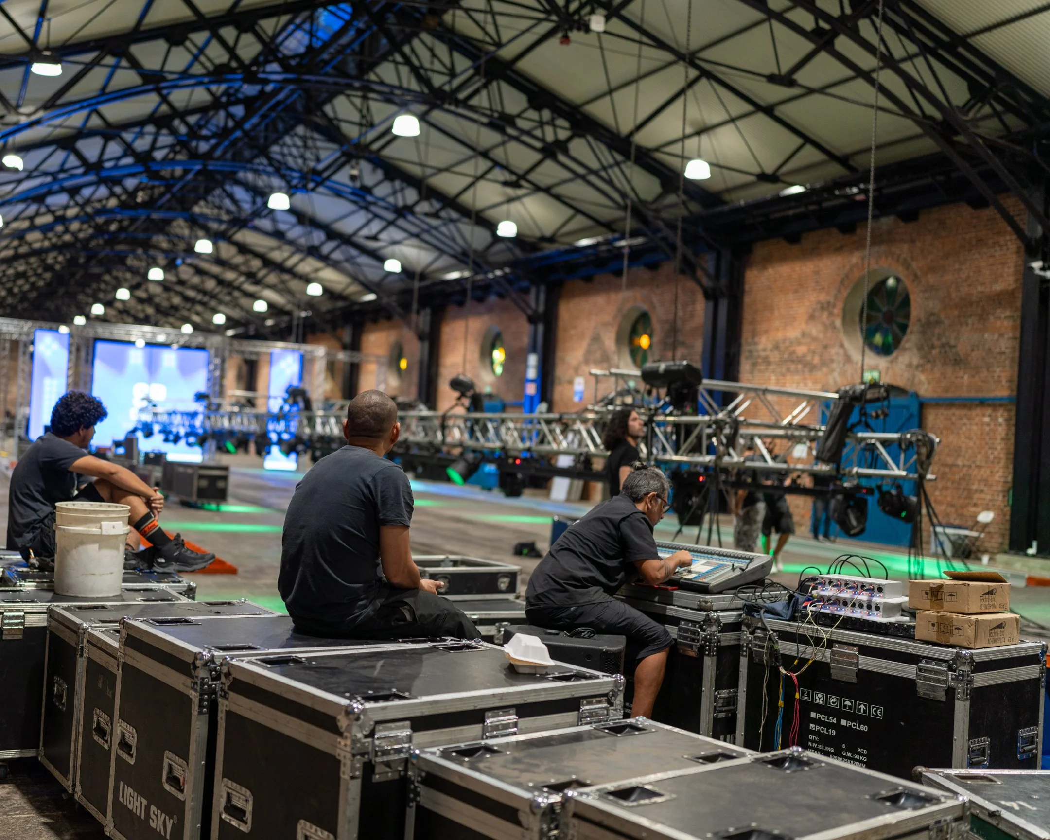 Personas preparando equipo técnico en un espacio amplio con estructura metálica y paredes de ladrillo, posible escenario para evento o concierto.