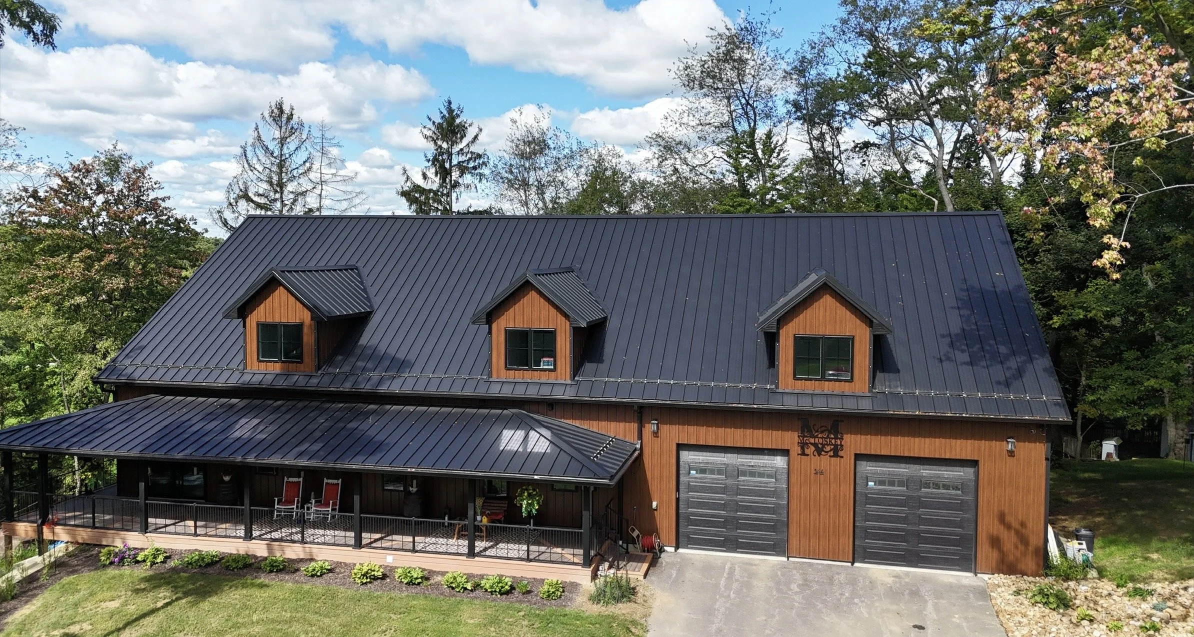 A two-story house with a black metal roof, wooden exterior, and three dormer windows on the upper floor, surrounded by green trees and a landscaped yard.