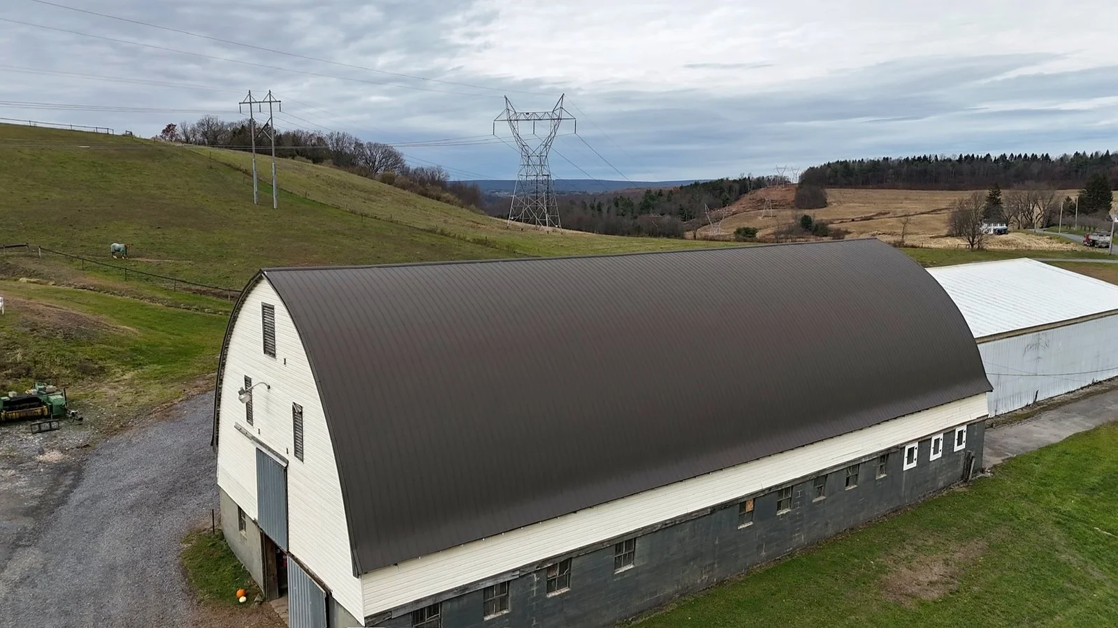 An aerial view of a large barn with a black curved roof and white walls, situated on a grassy farm with a gravel driveway. Rolling hills and power lines are visible in the background under cloudy skies.