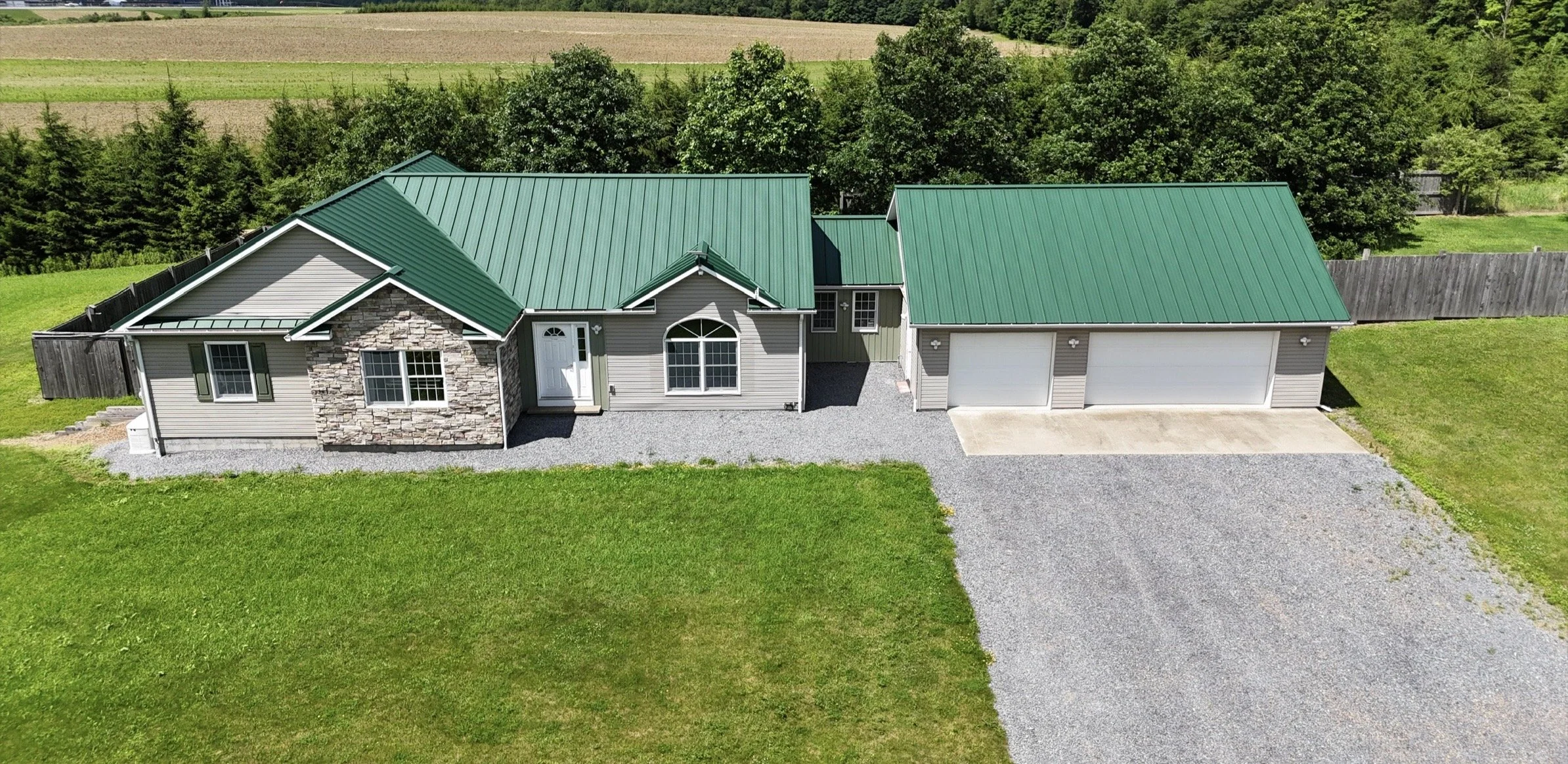 A house with a green metal roof, beige siding, and a stone accent wall, surrounded by a fenced yard and green grass, with farmland and trees in the background.