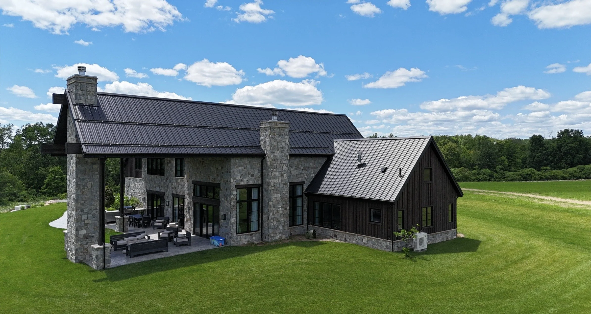 A modern house with stone and black siding exterior, featuring a metal roof, outdoor dining area, and surrounded by green grass and trees under a blue sky with scattered clouds.