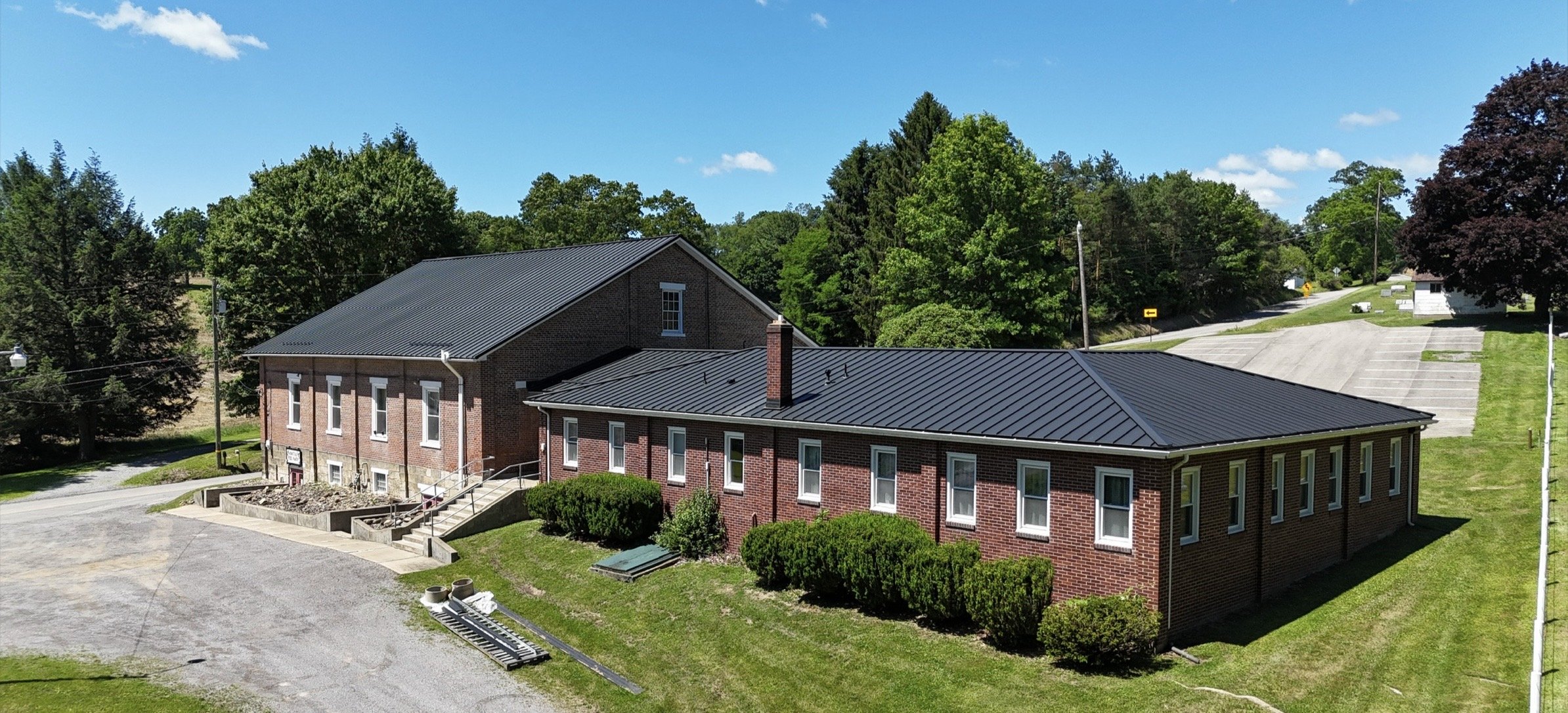 A red brick building with white windows and black metal roof in a rural area with trees and a parking lot.