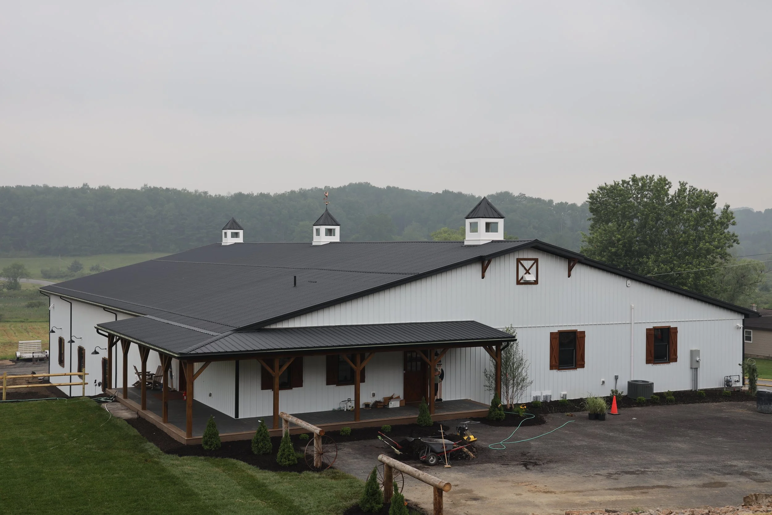 A modern white barn with a black metal roof and three small cupolas on top, situated in a rural area with green trees in the background. There is a backyard with a porch, some plants, and gardening equipment nearby.