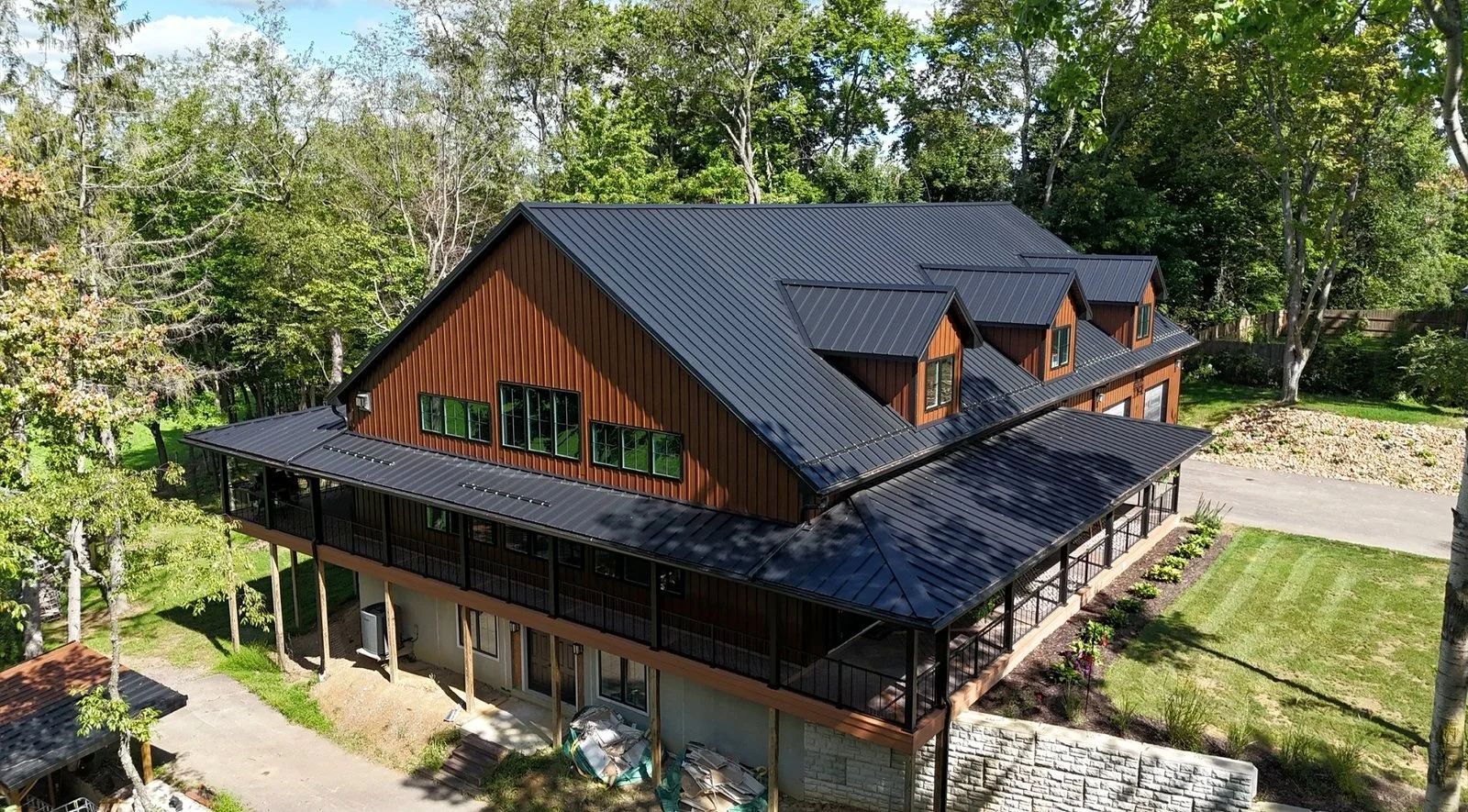 A large two-story house with brown vertical wooden siding and black metal roofing, surrounded by trees and a well-kept lawn, with a driveway on the right side.