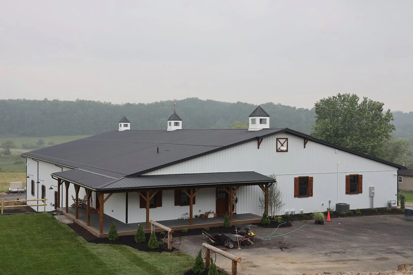 White barn with brown trim and a black metal roof, surrounded by a green lawn and a parking area, with trees and hills in the background on a cloudy day.