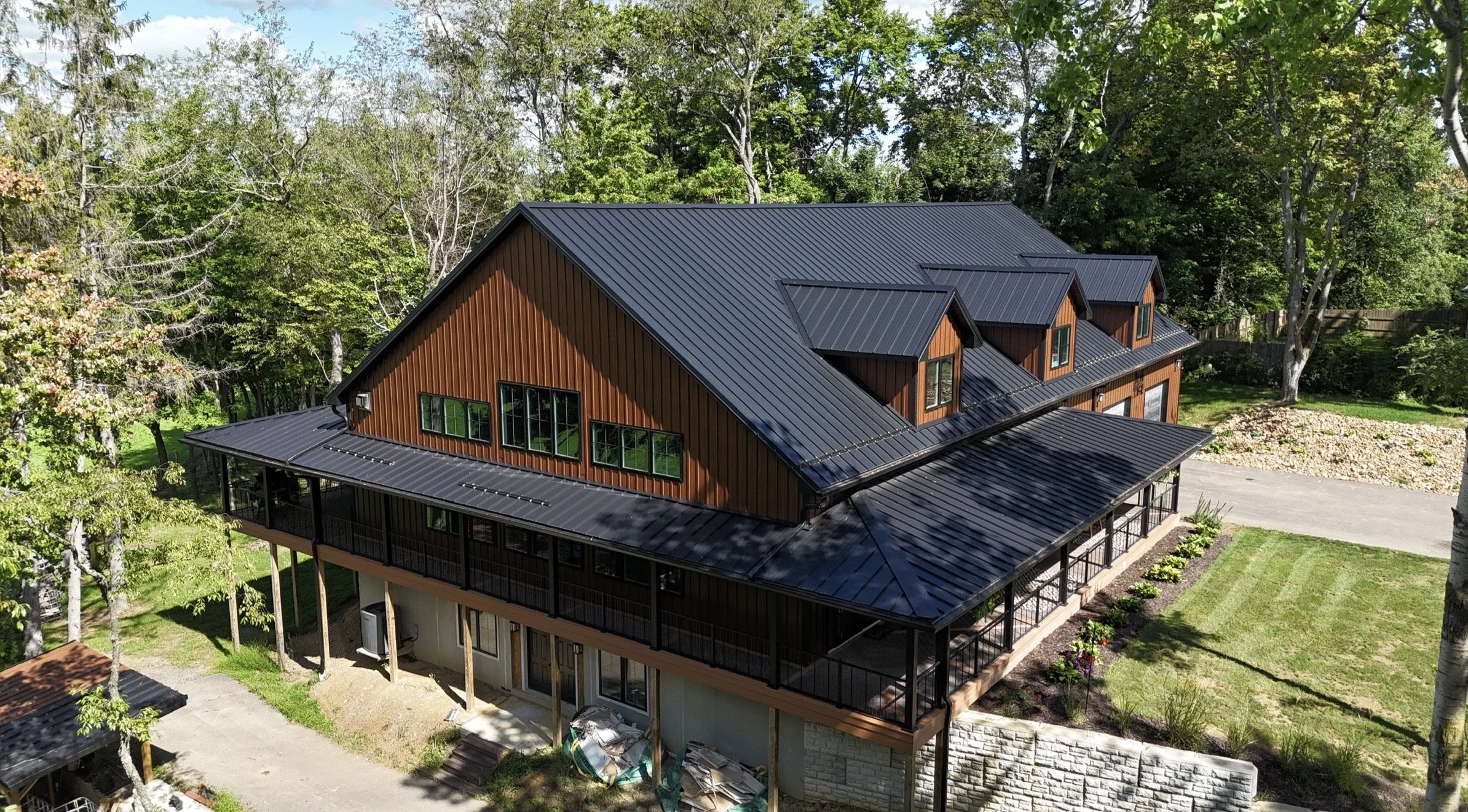 A modern two-story house with a black metal roof and brown wooden siding, surrounded by lush green trees and a manicured lawn.