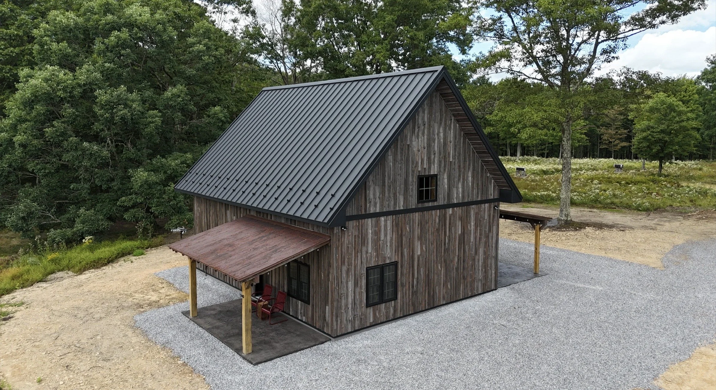 A wooden house with a steep black metal roof, set in a rural area with trees and greenery in the background.