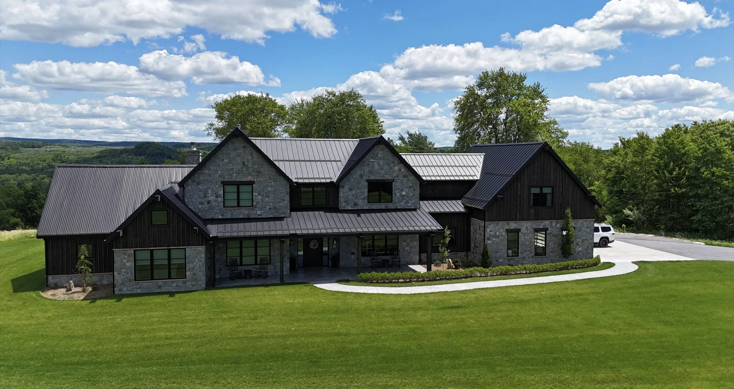 A modern two-story house with stone and dark wood siding, metal roof, large windows, front porch, surrounded by green lawn and trees, with a barbecue grill on the porch and a white SUV parked nearby, under a partly cloudy sky.