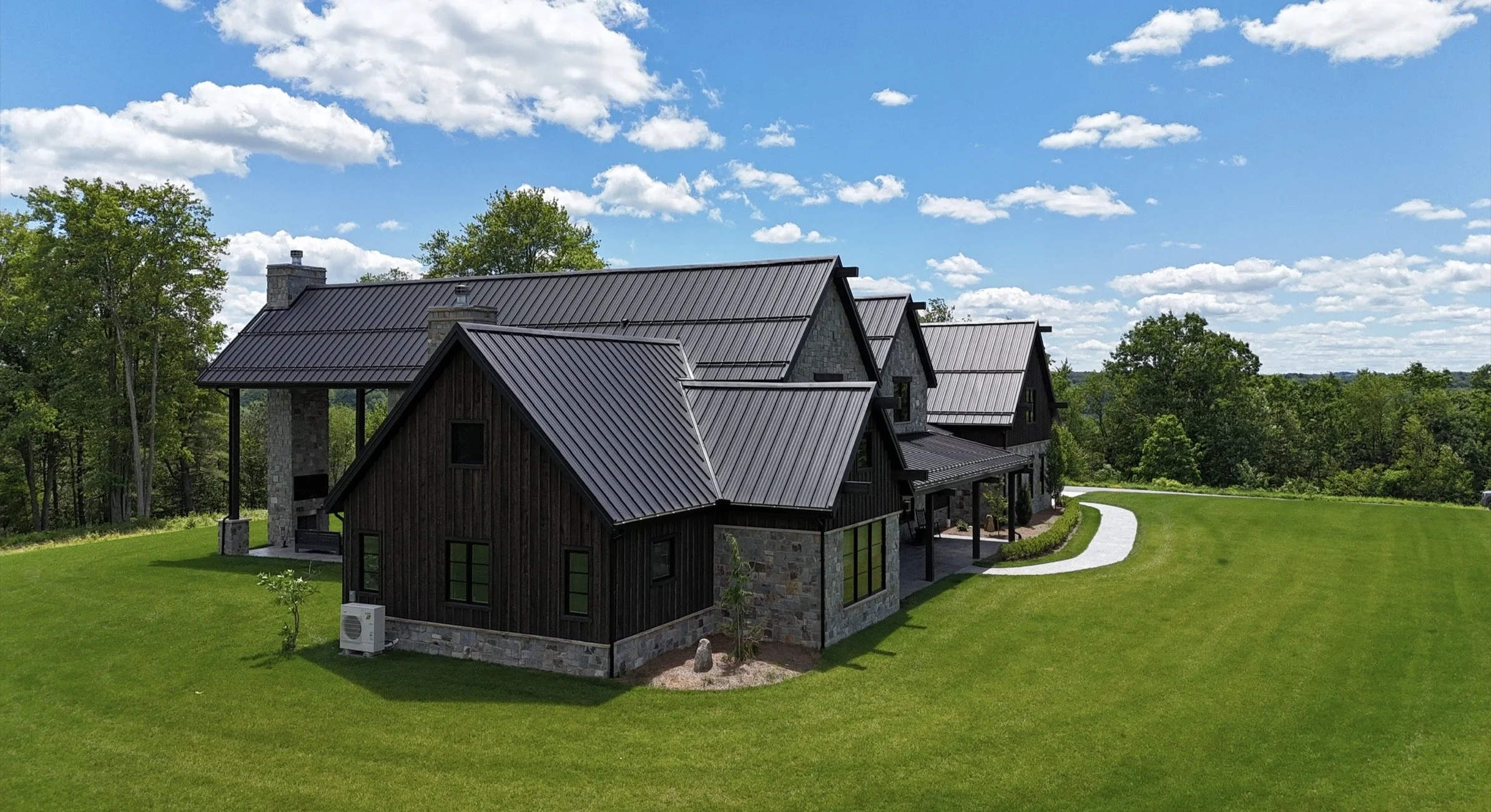 A large house with a black metal roof, stone and dark wood exterior, surrounded by a well-maintained green lawn and trees, with a curving concrete walkway and a bright blue sky with scattered clouds.