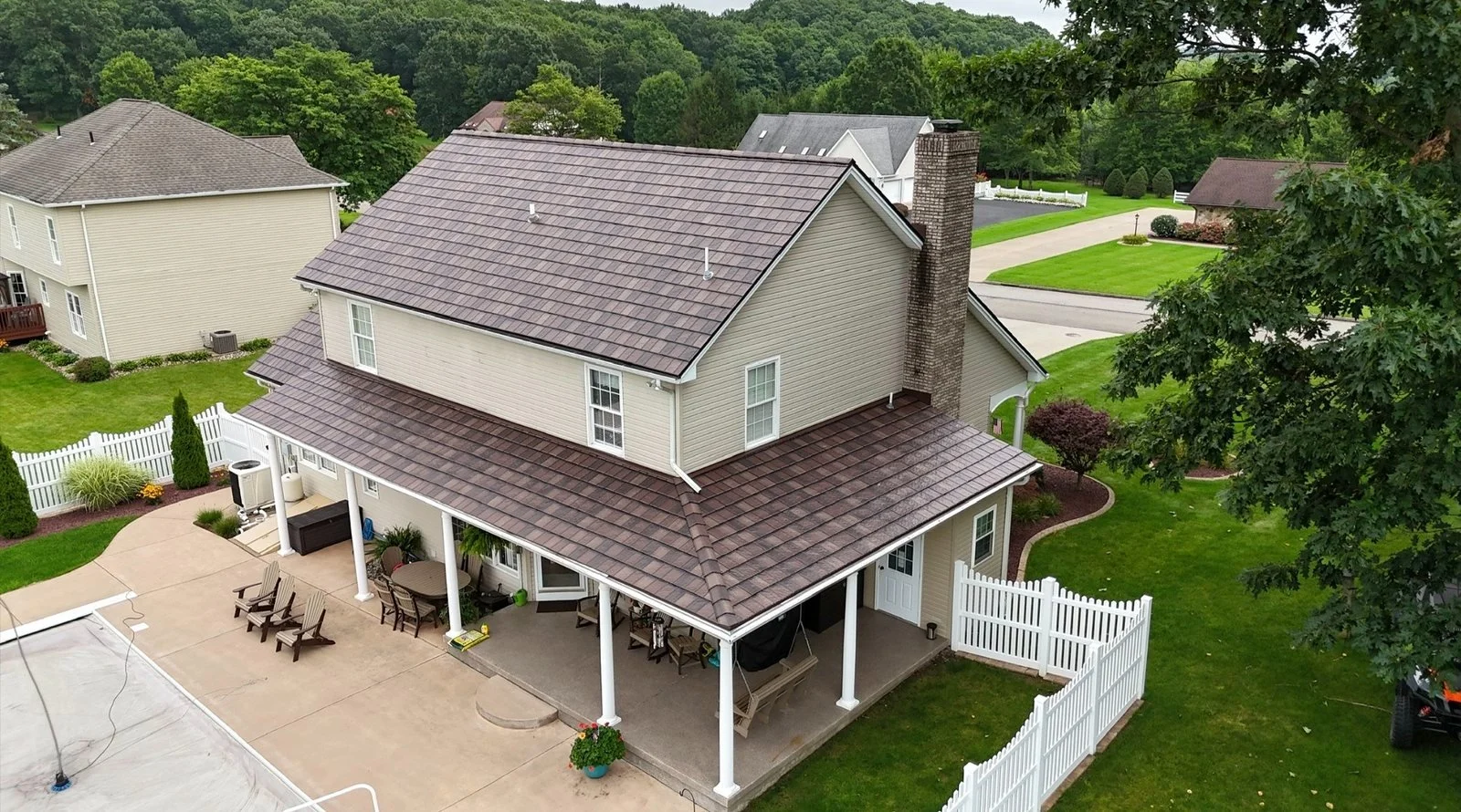 A two-story house with beige siding, a gray-tiled roof, and a brick chimney, surrounded by a well-maintained yard with green grass, trees, shrubs, and a white fence.