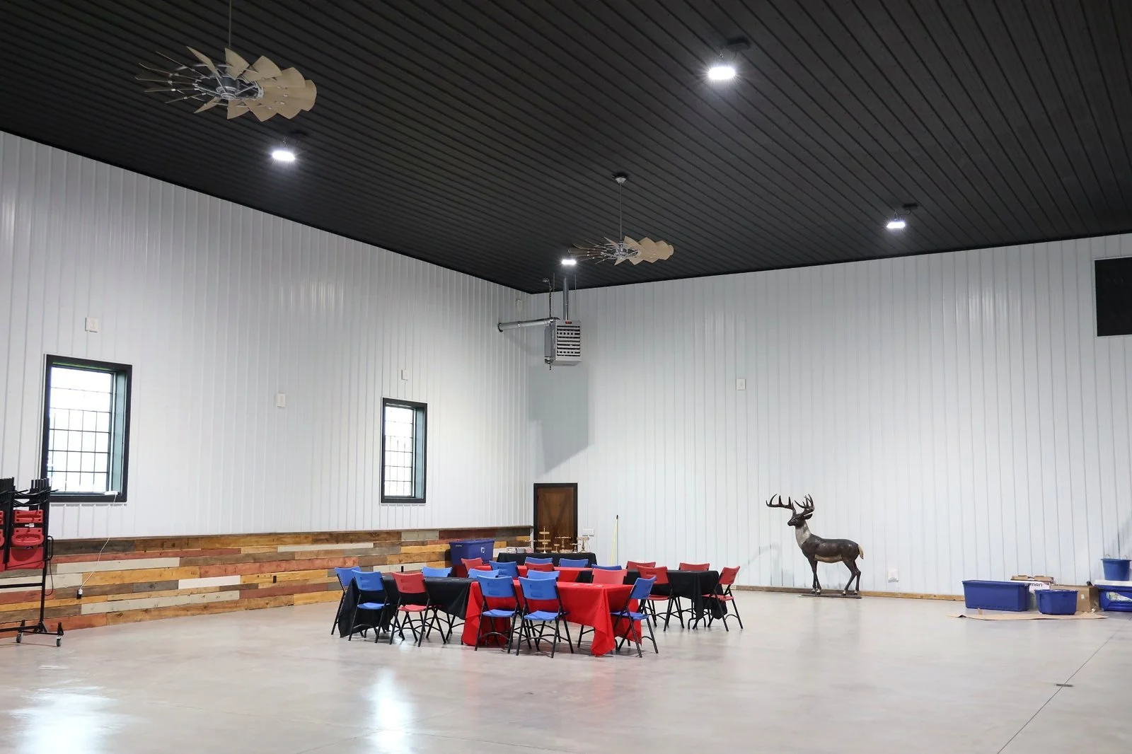 An indoor event space with black ceiling, white paneled walls, and a polished concrete floor. There is a group of tables with red and blue tablecloths and chairs, a rustic wooden step platform along the wall, a mounted deer statue, and scattered containers and items on the floor.