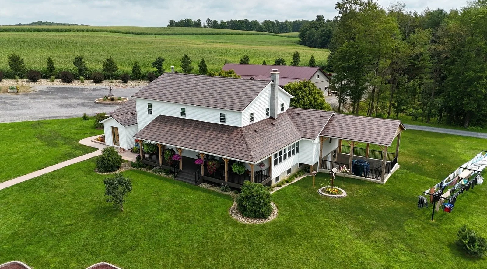 A large white house with a brown shingle roof, surrounded by a well-maintained lawn, trees, and a backdrop of distant green fields and forests.