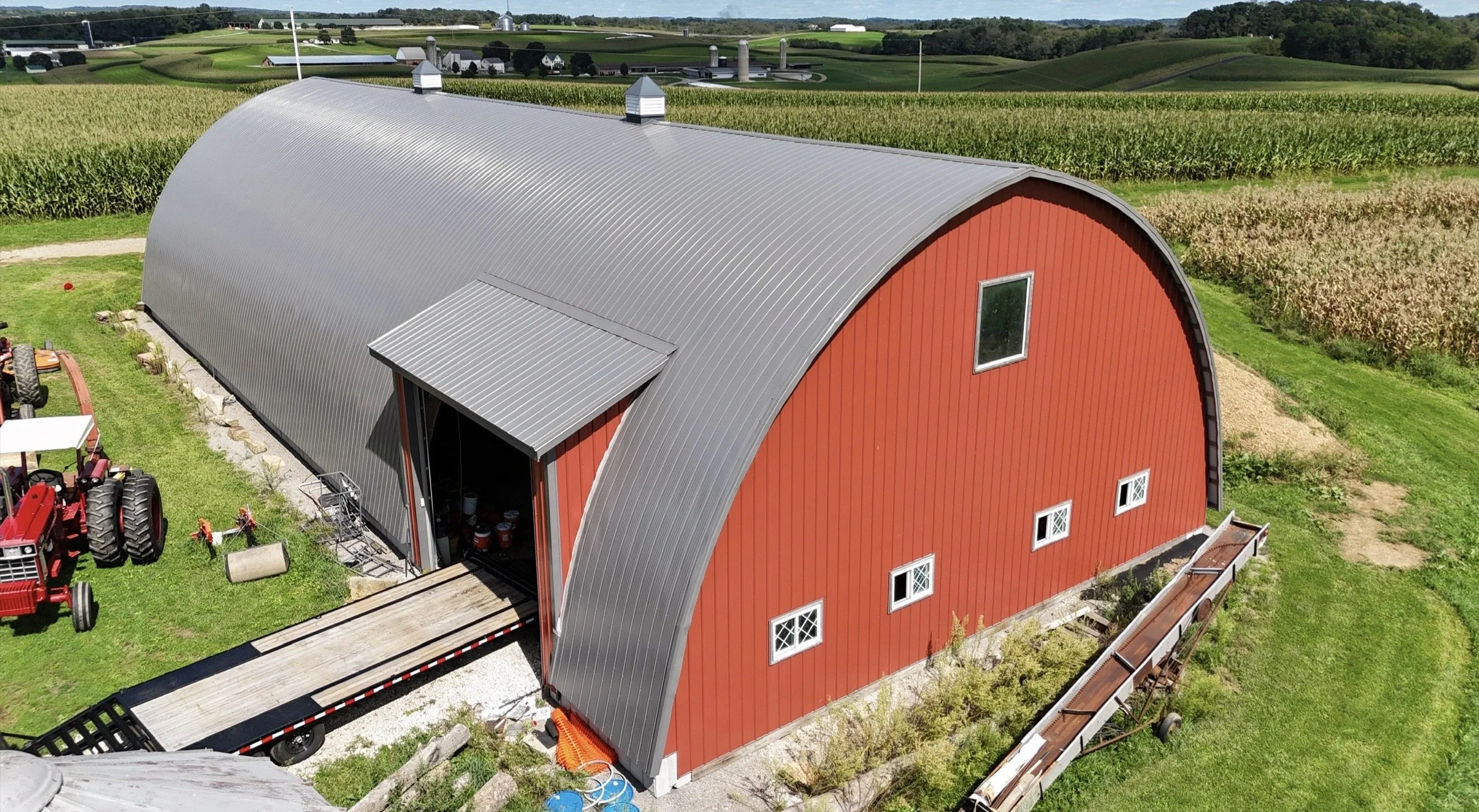 A red barn with a curved gray metal roof located in a rural farm setting, surrounded by farmland, trees, and hills in the background.