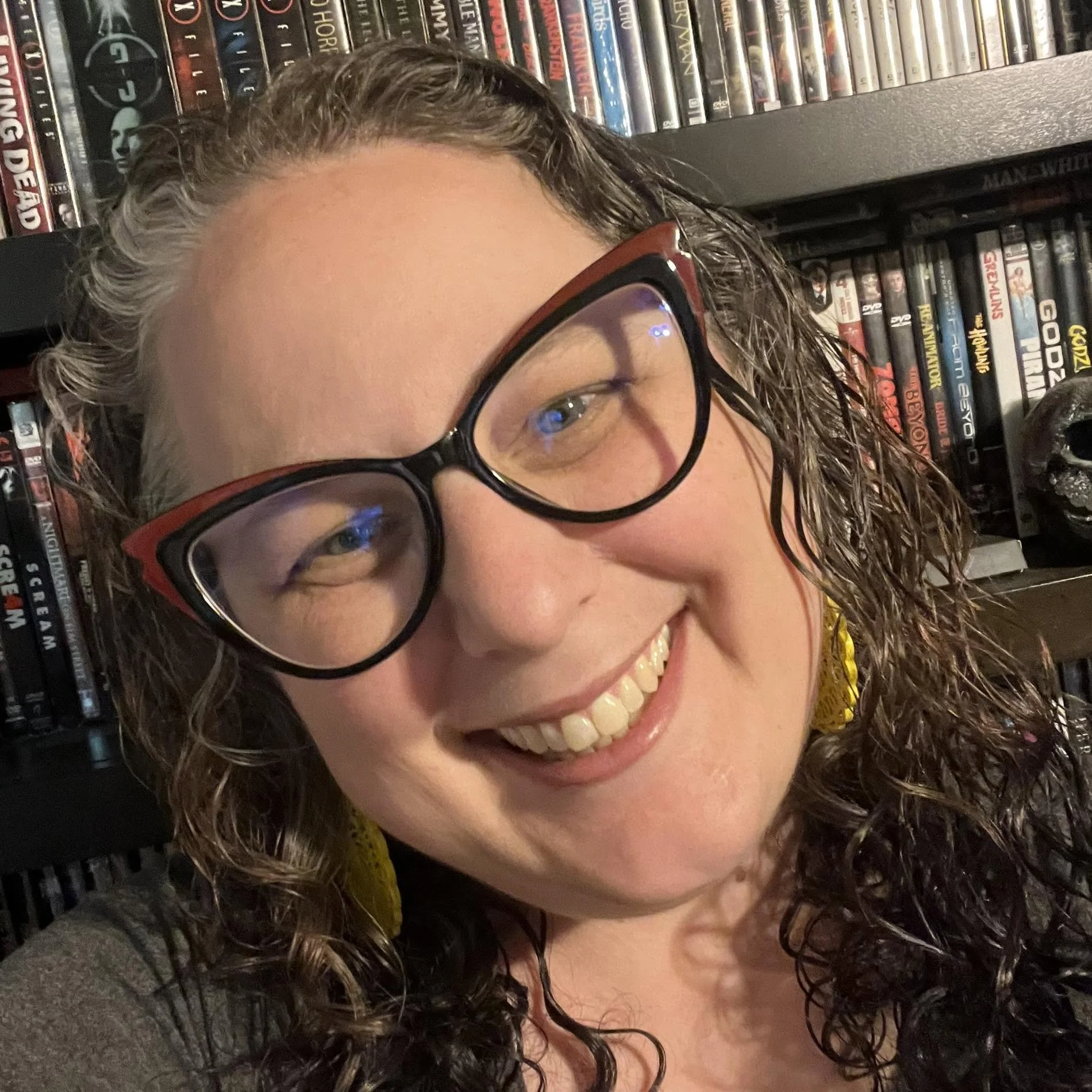 Close-up of a woman with curly hair wearing glasses, smiling, with a bookshelf filled with DVDs in the background.