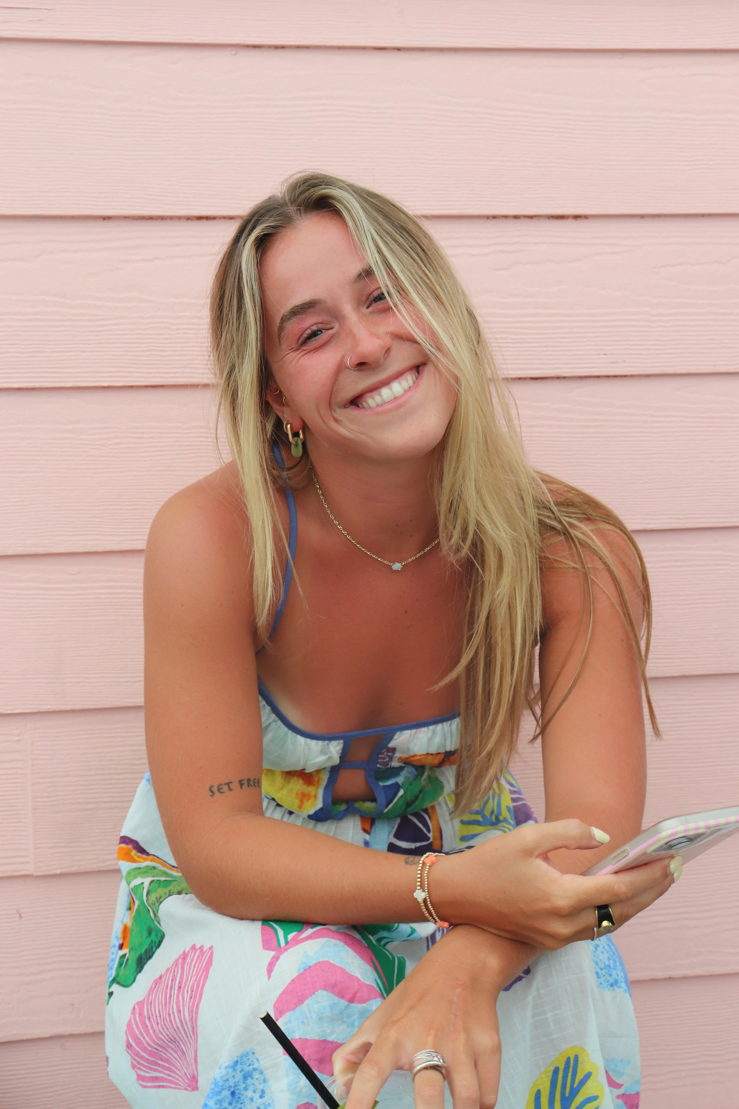 A smiling woman with long blonde hair, wearing a colorful dress with a floral pattern, earrings, and jewelry, sitting in front of a pink wooden wall, holding a smartphone.