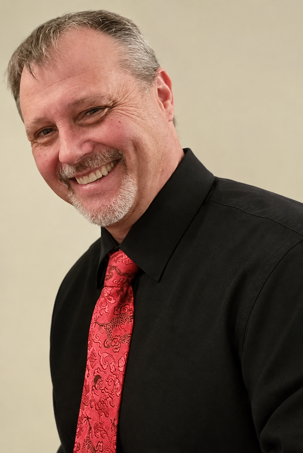 Smiling middle-aged man with a gray goatee wearing a black shirt and red paisley tie