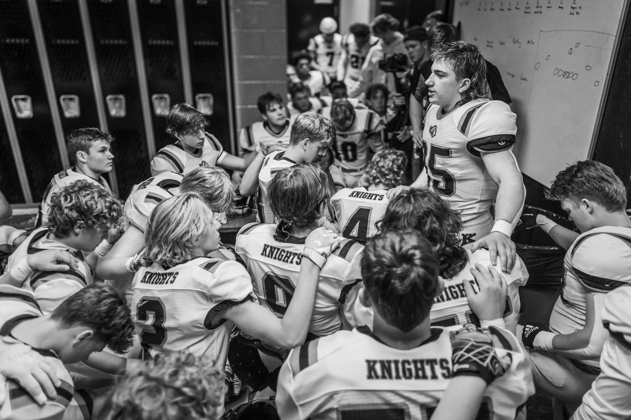 Youth football team huddled in a locker room as a player addresses them beside a whiteboard