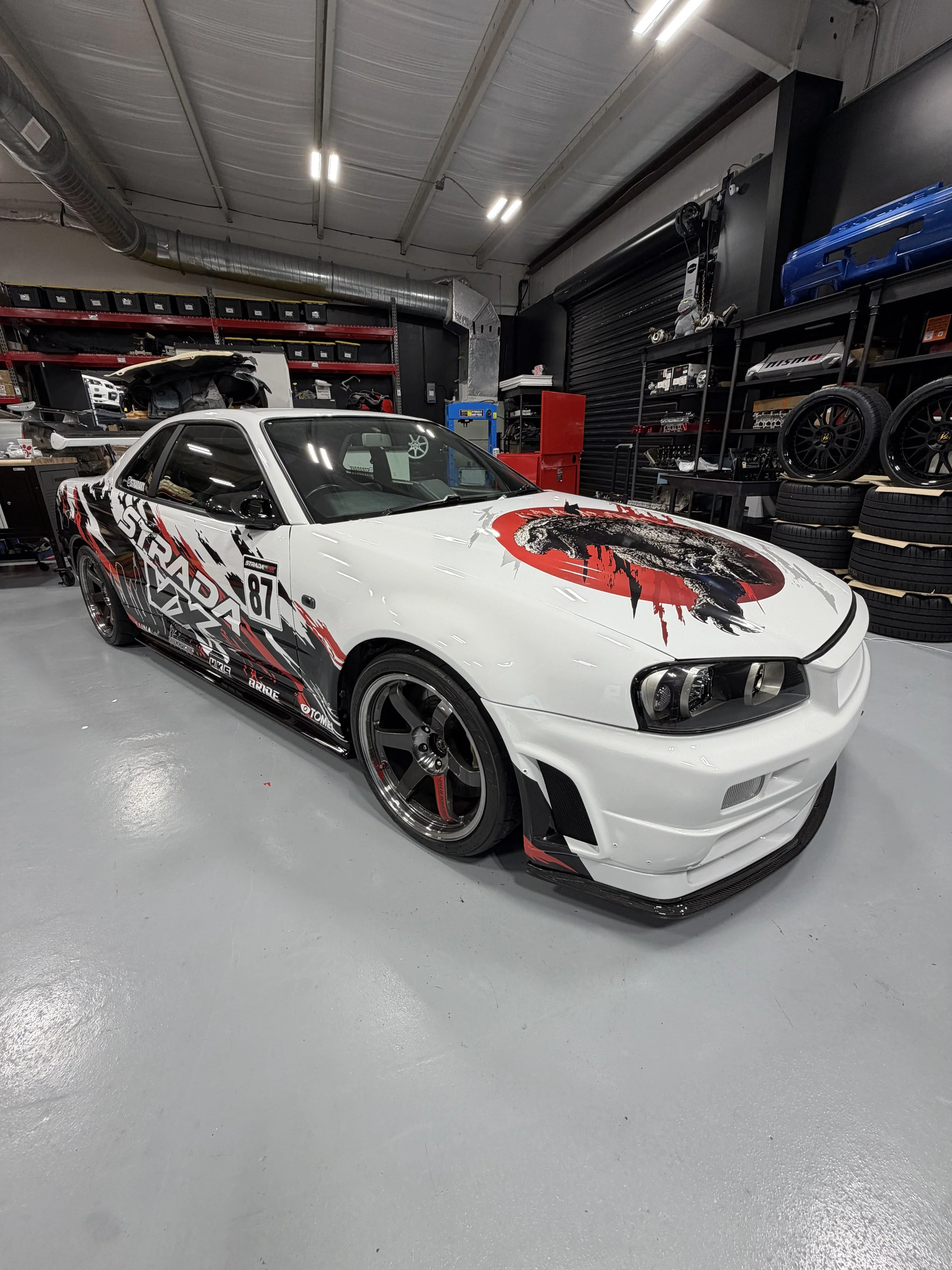 Modified white sports car with bold red-and-black graphics parked inside an automotive workshop surrounded by tools, tires, and parts.
