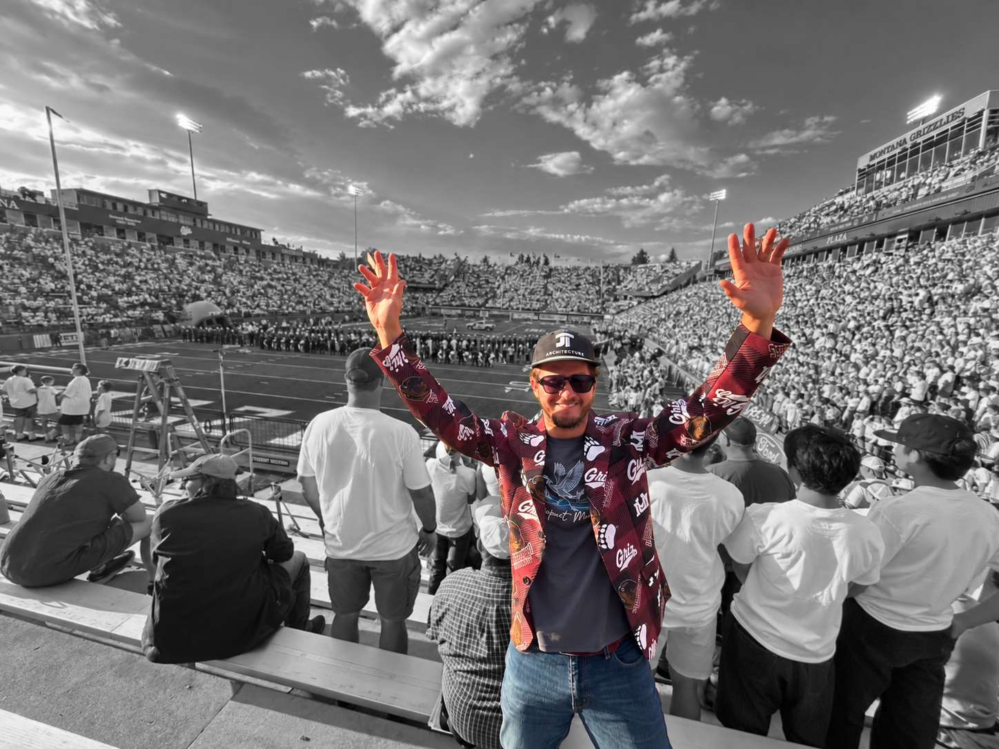 A  local Missoula man wearing sunglasses, a cap, and a maroon jacket with University of Montana Griz Logos, smiling and raising his arms in celebration at a crowded football stadium.
