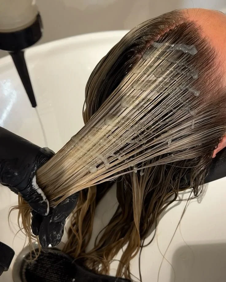 A person receiving hair treatment with sections of their wet hair being coated with a product, in a salon sink view.