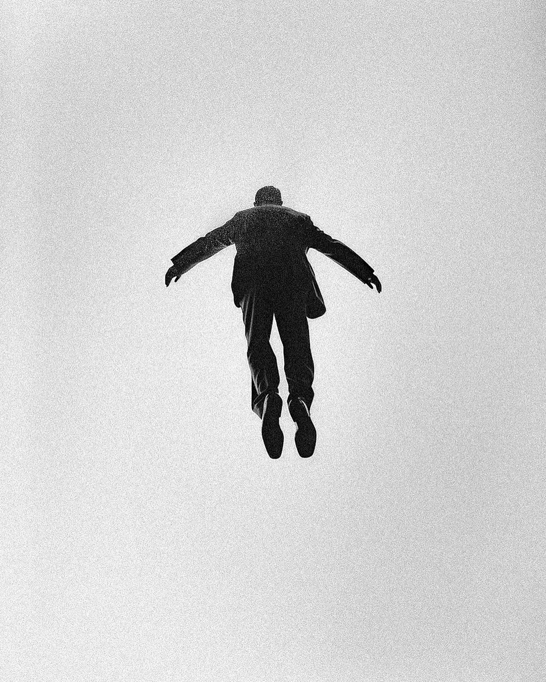Silhouette of a man in a suit seemingly floating or flying in the air with arms outstretched against a plain background.