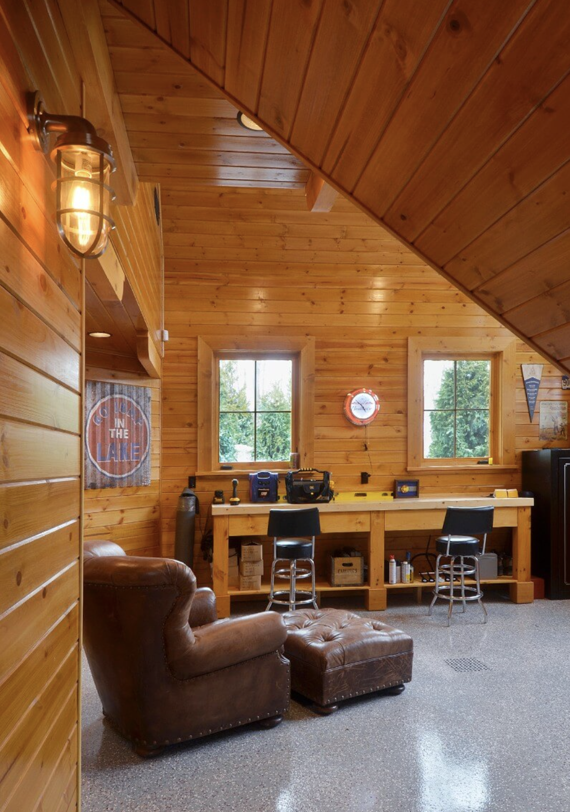 A cozy wood-paneled room with a sloped ceiling, featuring a small workspace with a long wooden desk, two chairs, and various tools and equipment. There are two windows with a view of greenery outside, a leather armchair with an ottoman, and a wall cl