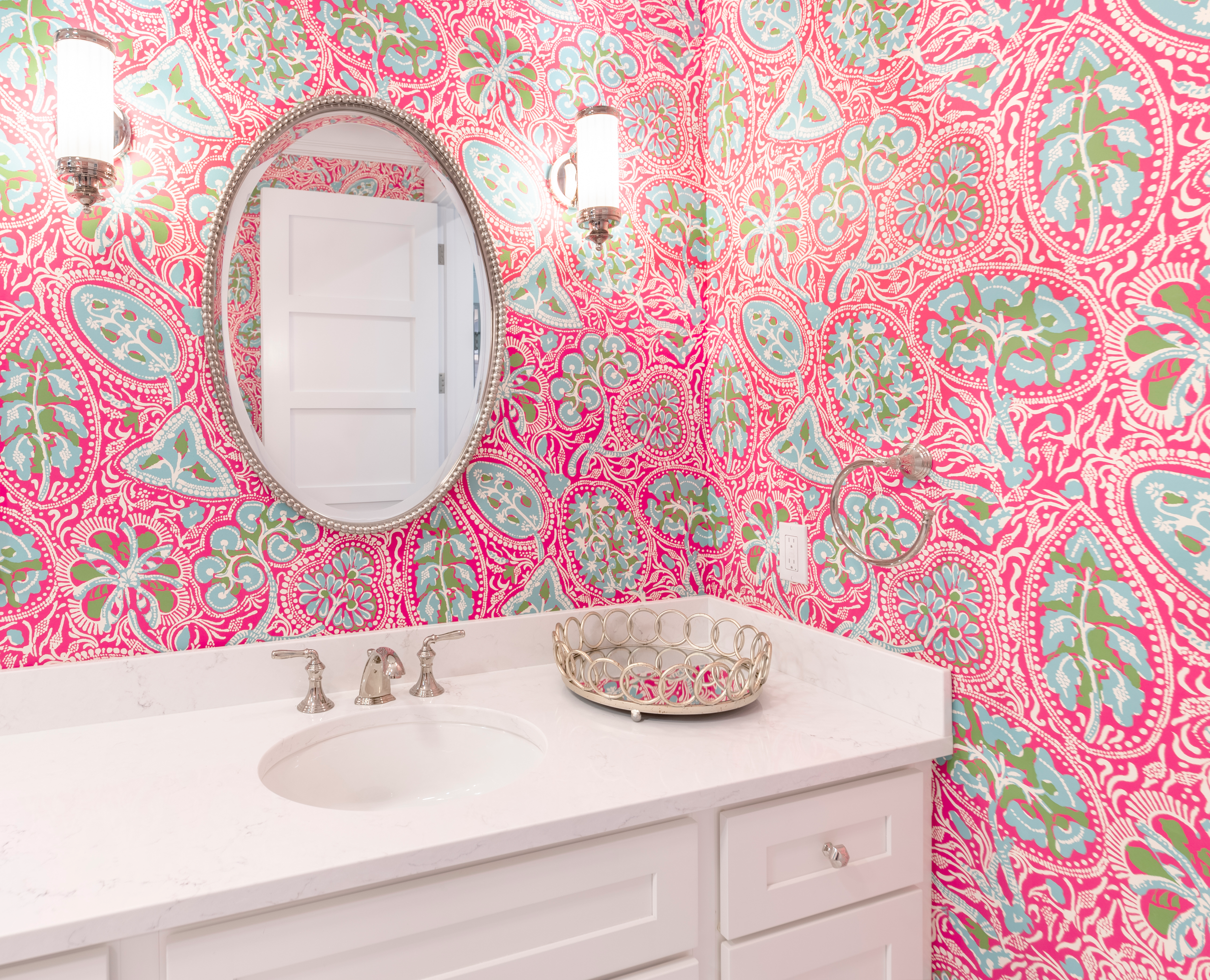 Bathroom with a pink, green, and blue floral patterned wallpaper, a white vanity with a marble countertop, a small oval mirror with a beaded frame, two wall-mounted light fixtures, and a gold wire basket on the countertop.