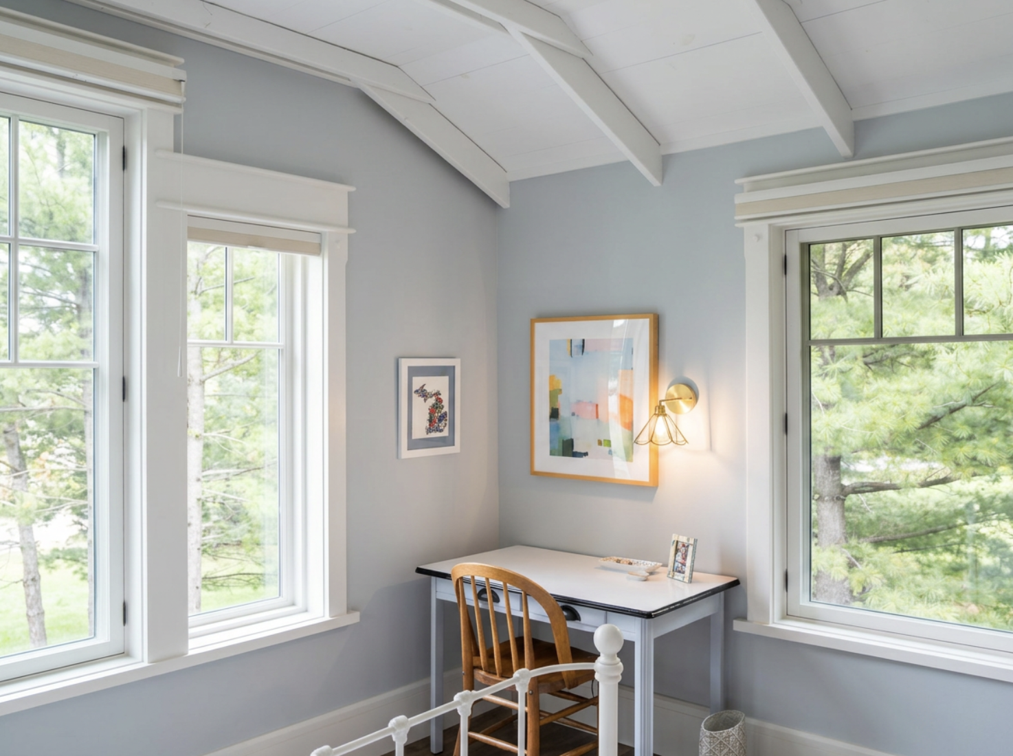 Corner of a cozy bedroom with two large windows showing green trees outside, light blue walls, a white desk with a wooden chair, and framed abstract art on the walls, illuminated by natural light and a small wall sconce
