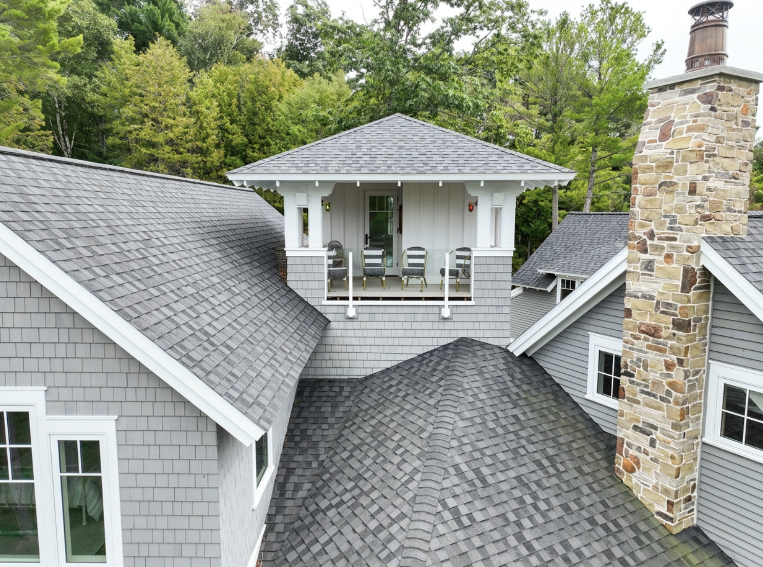 Aerial view of a house with multiple gray shingle roofs, a stone chimney, and a small upper balcony with chairs, surrounded by green trees.