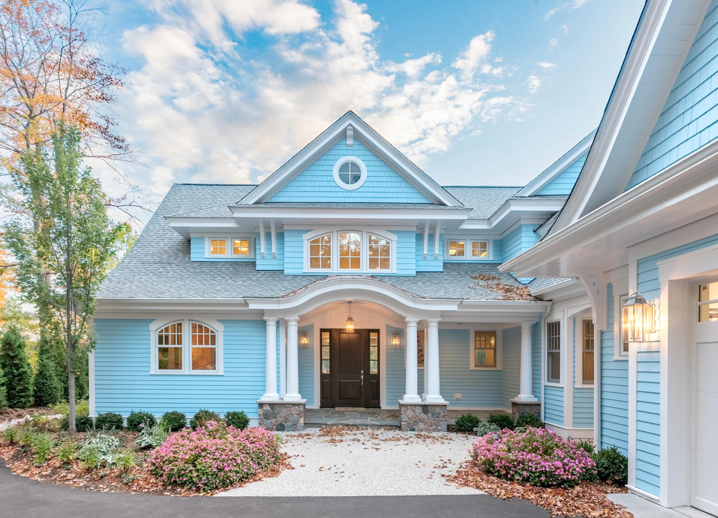 Front view of a blue house with a gabled roof, white trim, stone pillars at the entrance, and surrounded by trees and pink flowers, with a partly cloudy sky in the background.
