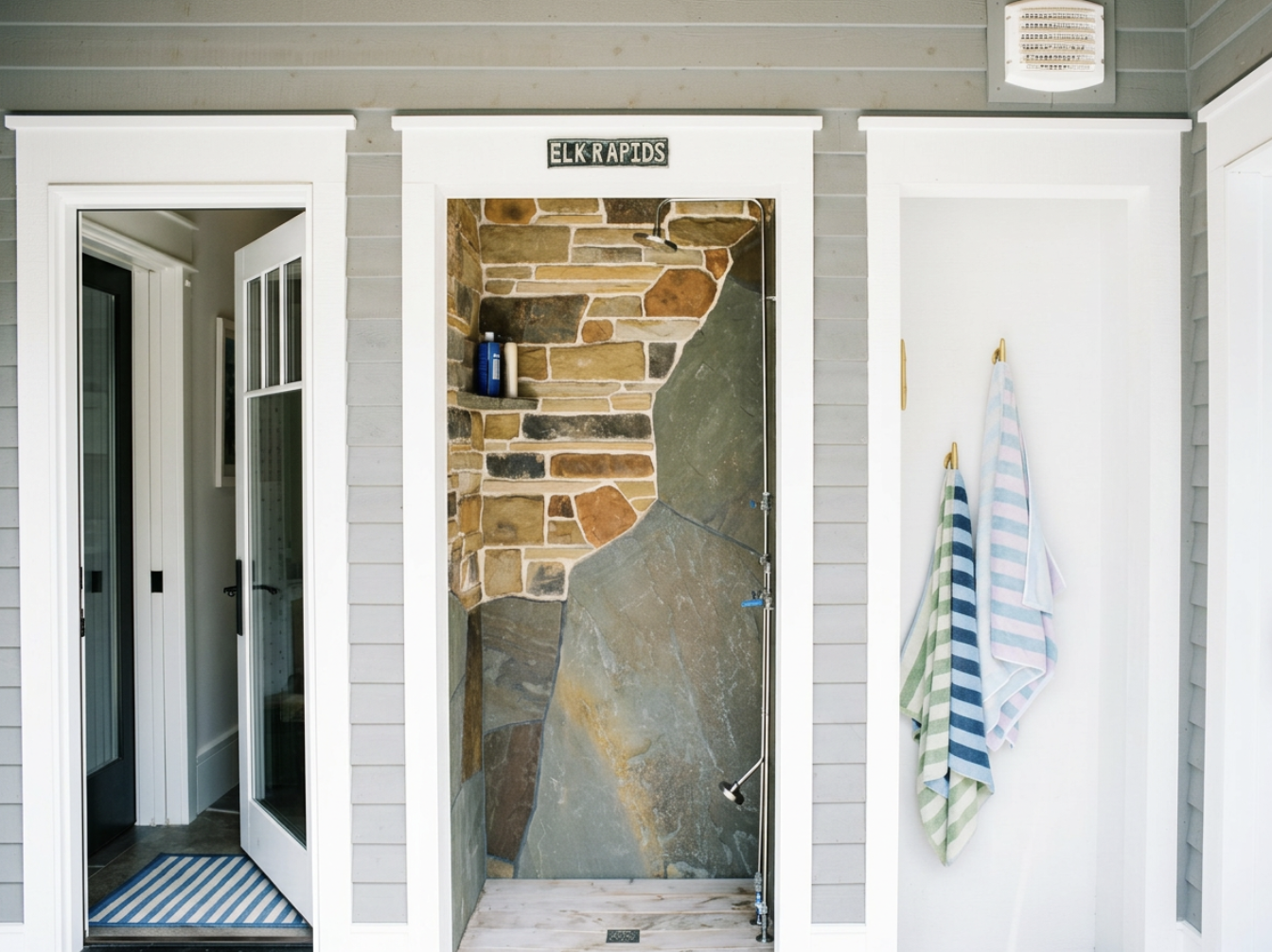 Outdoor shower with brick, stone, and tile walls, labeled with 'Elk Rapids' sign, next to a white door with striped towels hanging.