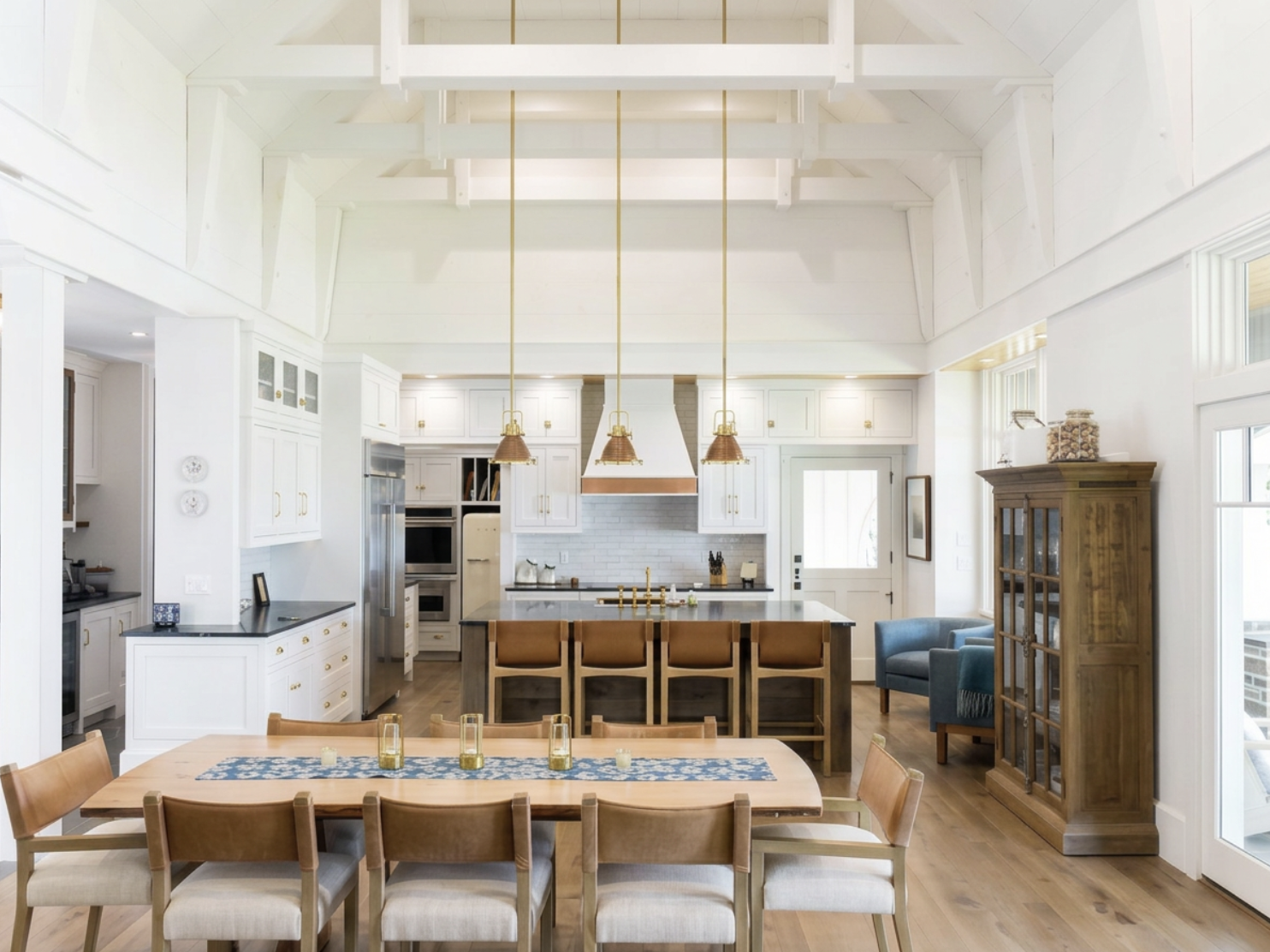 Bright kitchen with white cabinets, a large island with wooden chairs, a dining table with wooden chairs, and a glass-front cabinet. The kitchen has a vaulted ceiling with exposed beams, hardwood floors, and large windows.