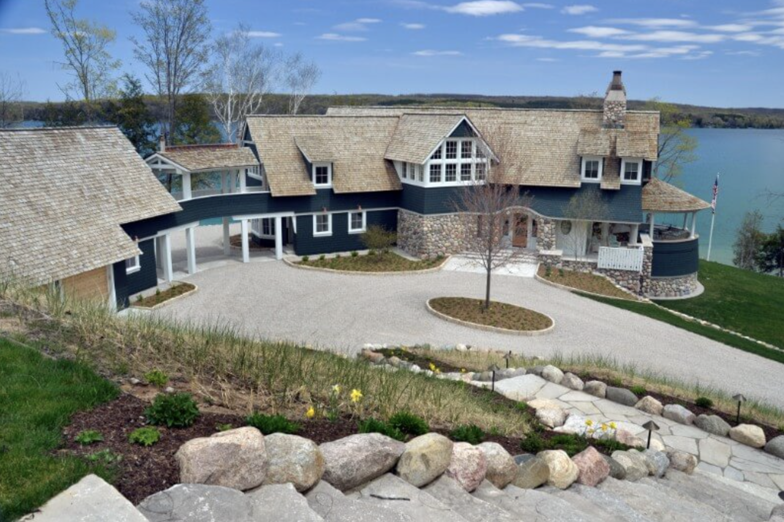 Large lakeside house with a mix of stone and dark siding, multiple gabled roofs, surrounded by greenery and a water body in the background.