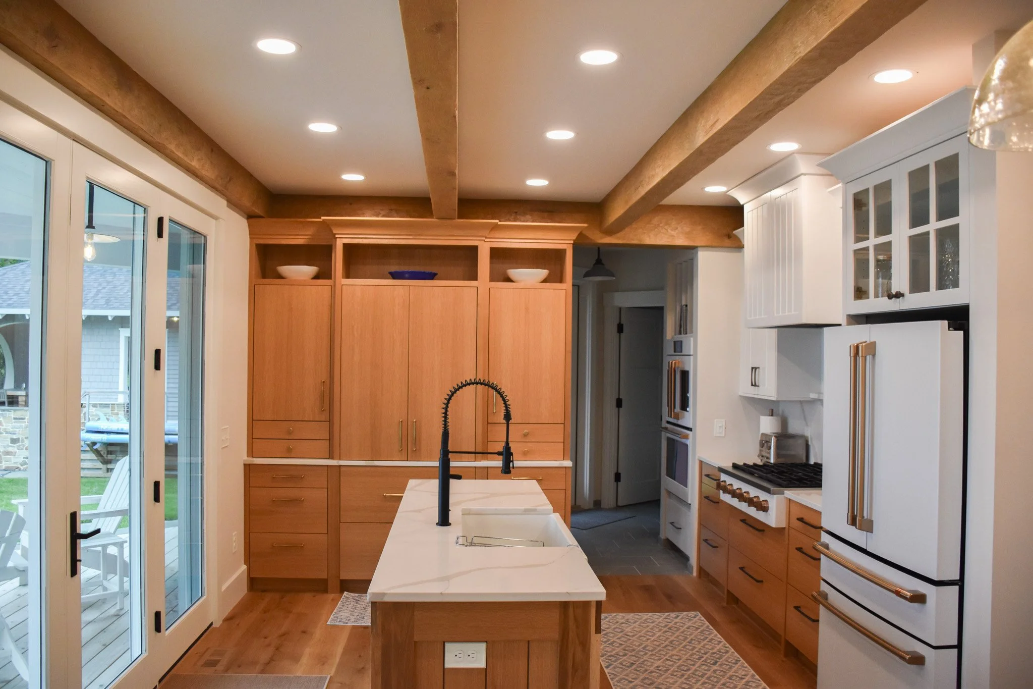 A modern kitchen with white cabinets, wooden accents, and a central island with a black faucet. Double glass doors lead outside, and recessed ceiling lights illuminate the space.