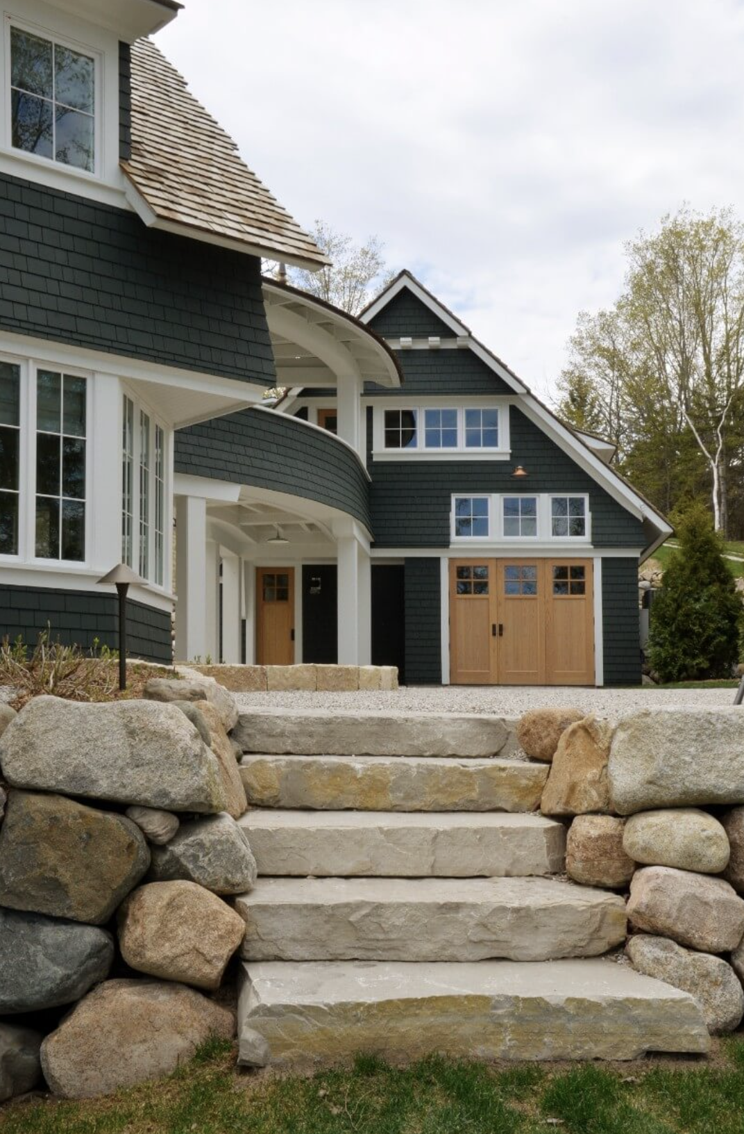 Front view of a modern house with dark blue siding, wooden garage doors, and large white-framed windows, accessed by stone steps with stacked rocks on either side.
