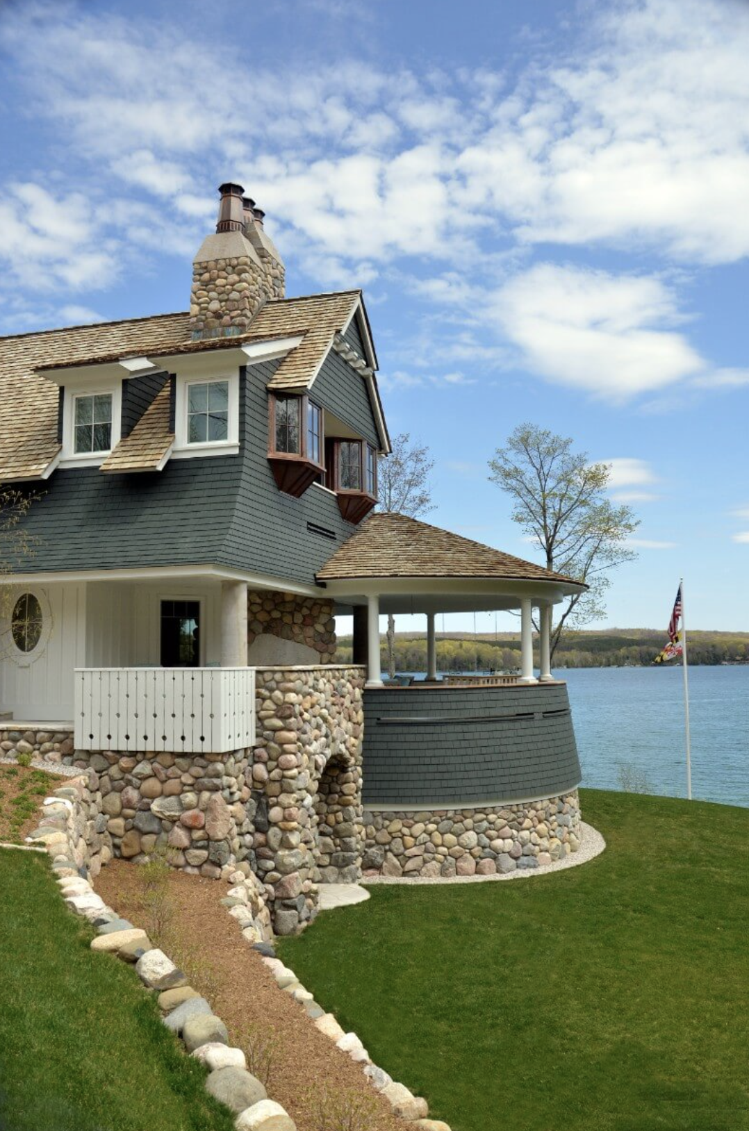 A house by a body of water with a stone and wooden exterior, a covered patio, a flagpole, trees, and a partly cloudy sky.